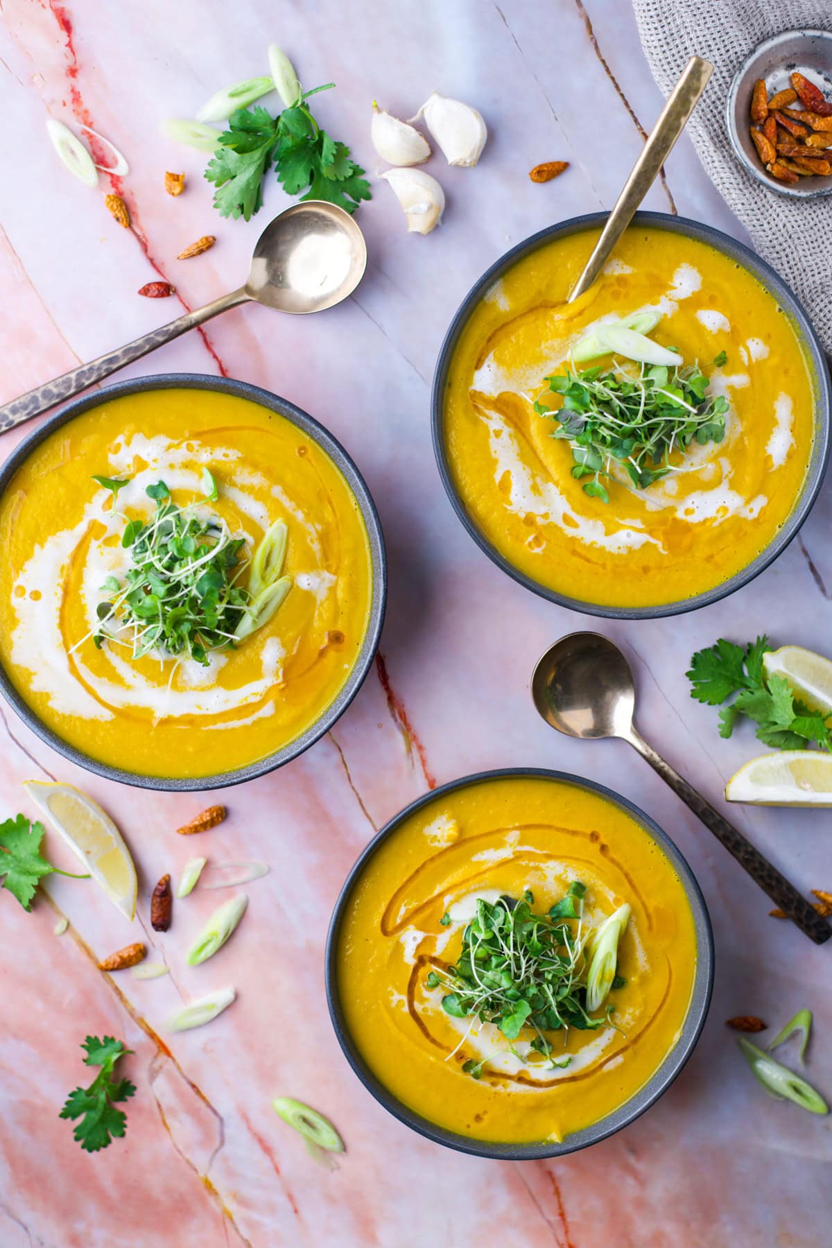 Three bowls of creamy yellow soup garnished with microgreens, sliced green onions, and a swirl of cream, placed on a marble surface with spoons and fresh herbs, lemon wedges, and garlic cloves nearby.