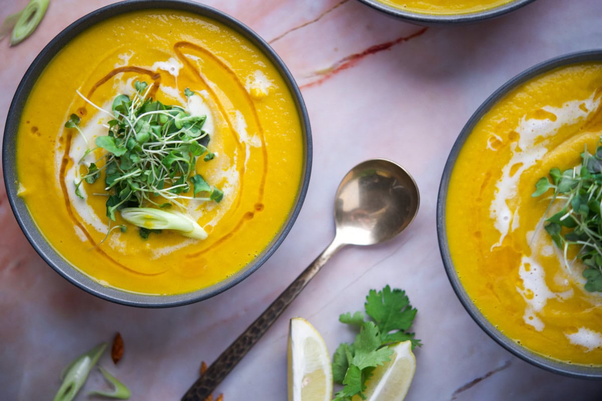 A bowl of creamy yellow soup topped with microgreens, sliced green onions, and a swirl of cream, next to a spoon, lemon wedges, and cilantro on a light pink marble surface.