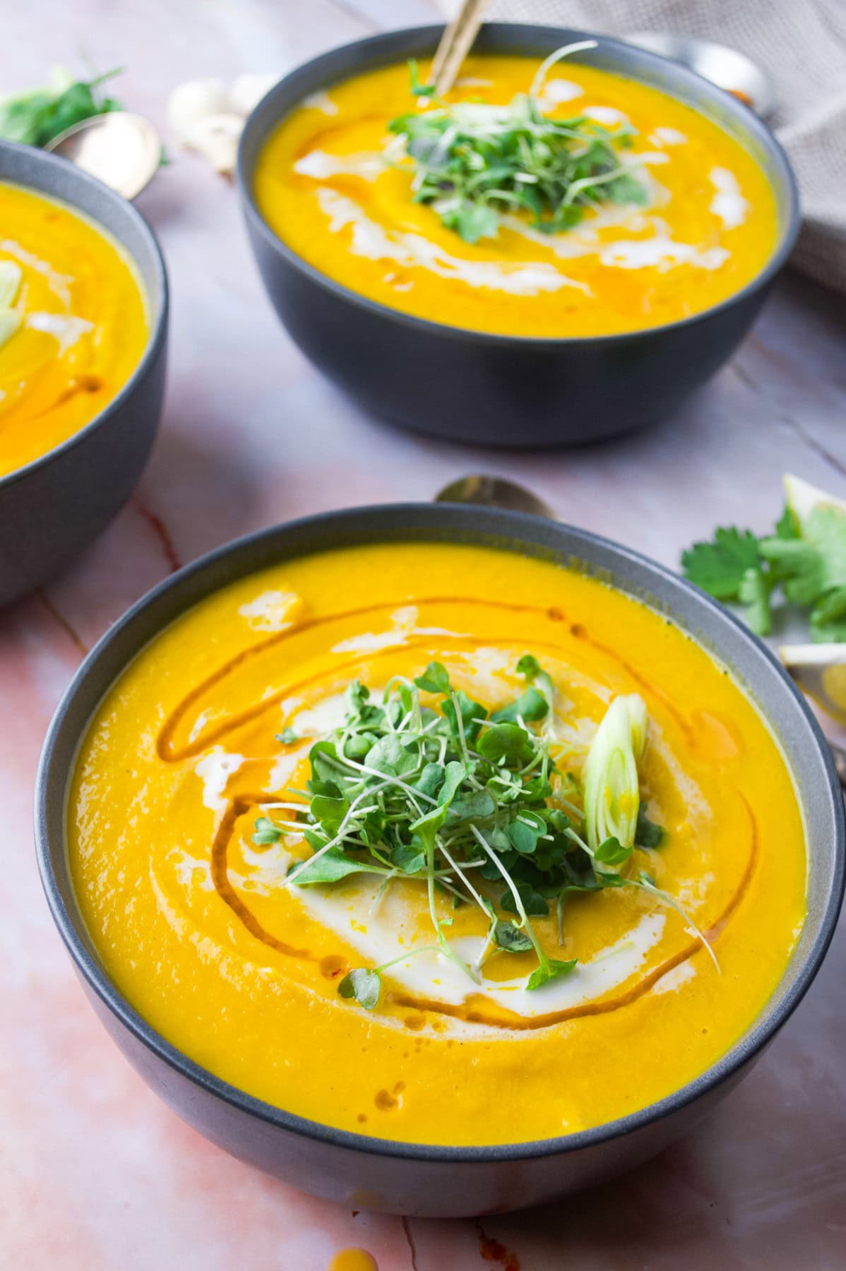 Three bowls of Thai Red Curry Butternut Squash Soup, garnished with a swirl of cream, microgreens, and a drizzle of oil, sit on a marble surface. One bowl is in the foreground and two are slightly blurred in the background.
