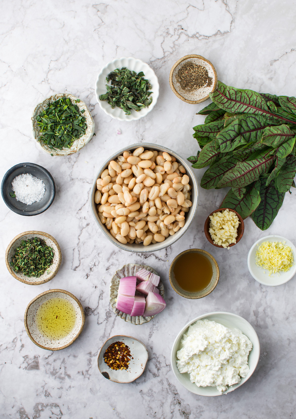A variety of small bowls on a marble surface containing white beans, fresh herbs, leafy greens, salt, pepper, olive oil, vinegar, minced garlic, shallot, zest, red pepper flakes, and ricotta cheese. White Bean and Sorrel Dip