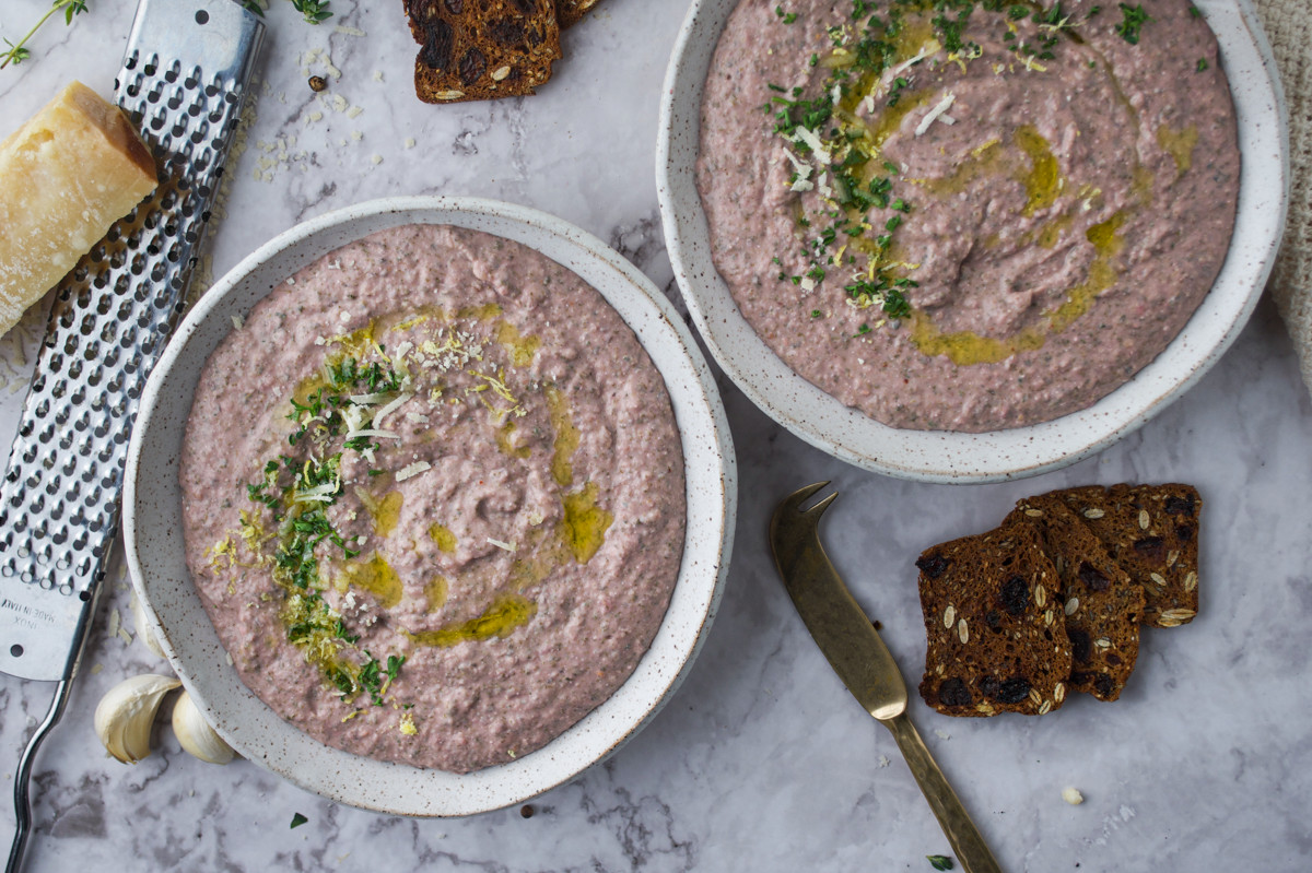 Two bowls of creamy dip garnished with herbs, parmesan, and olive oil sit on a marble surface, surrounded by crisp bread slices, a wedge of cheese, garlic, and a grater.