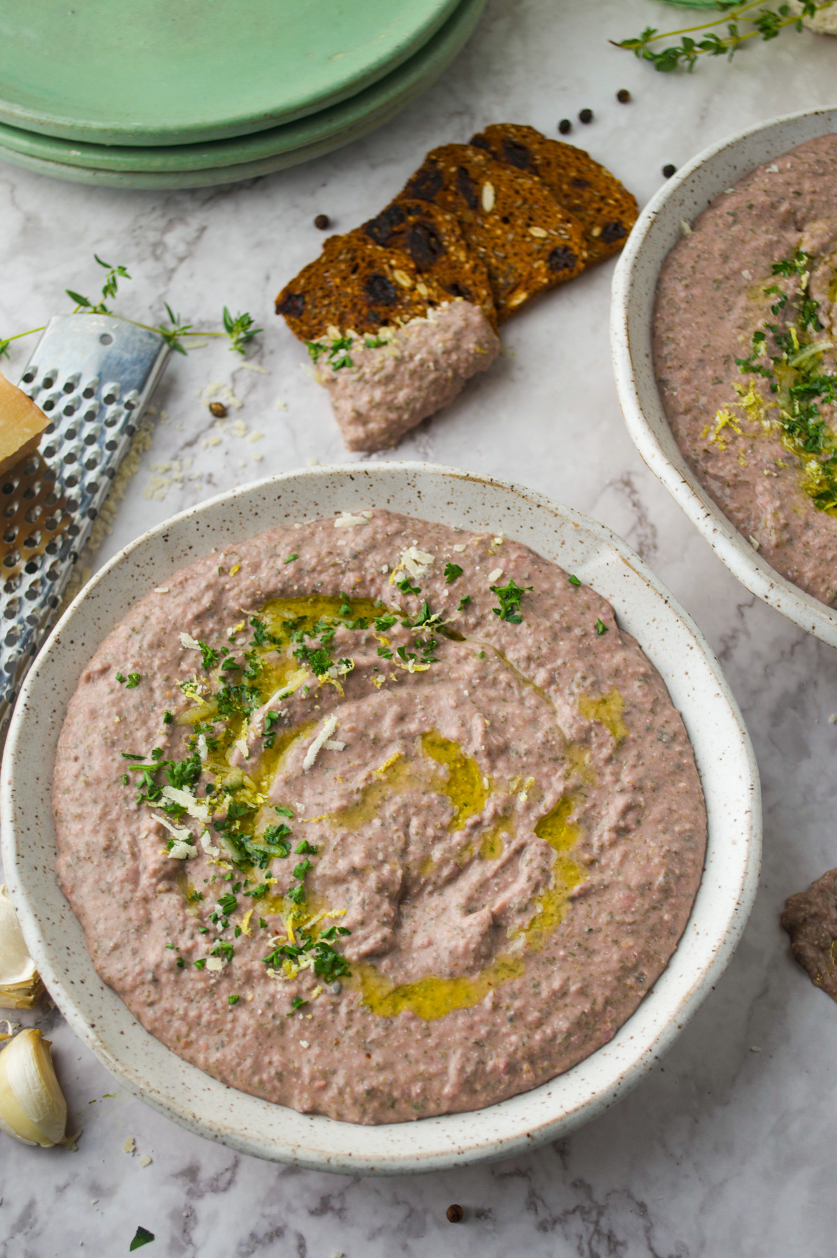 A bowl of creamy dip garnished with chopped herbs and olive oil, with crackers and fresh garlic nearby on a marble surface.
