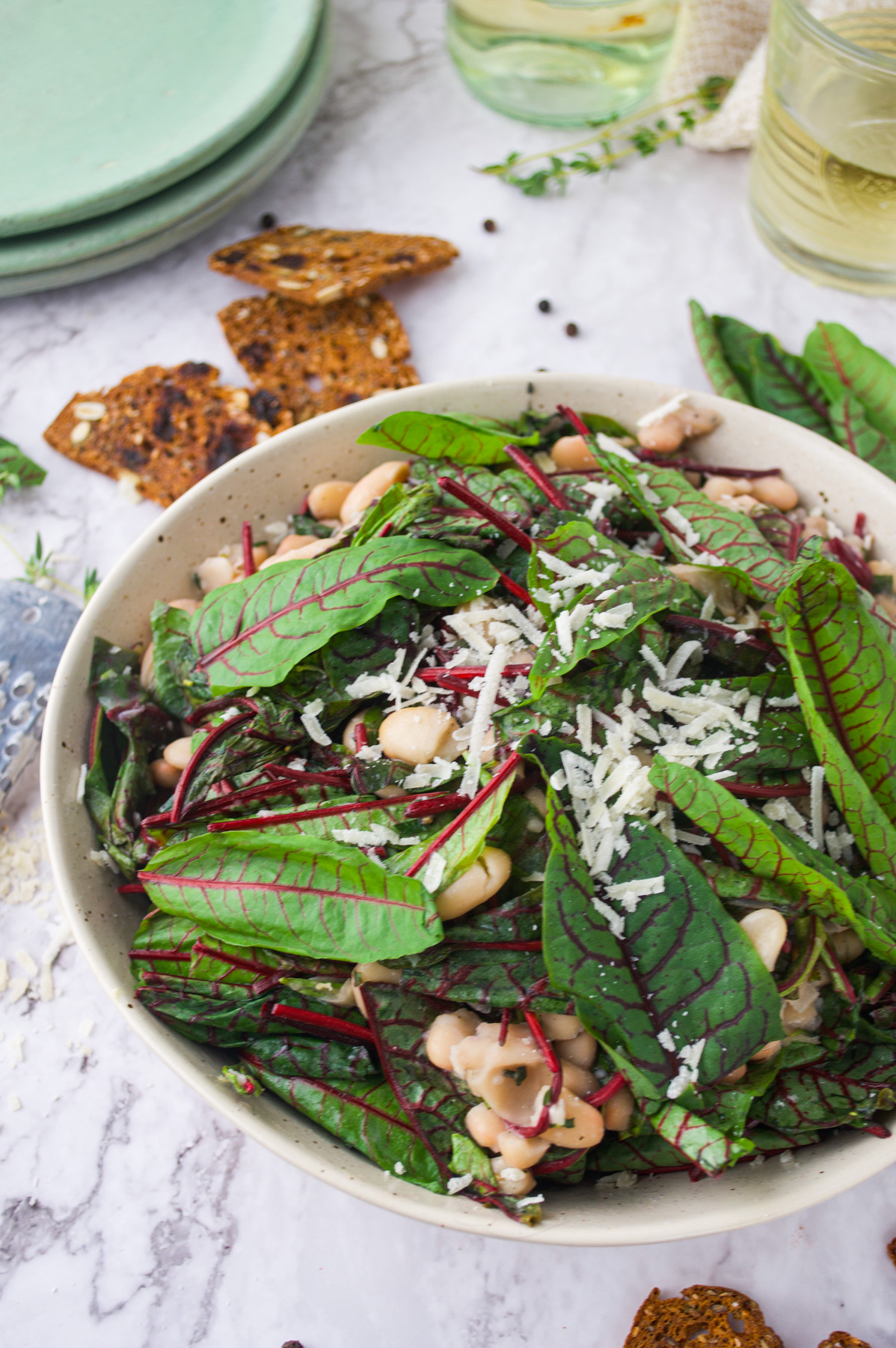 A bowl of fresh salad with leafy greens, white beans, and shredded cheese on top, placed on a marble surface with crackers, green plates, and a glass of white wine nearby.