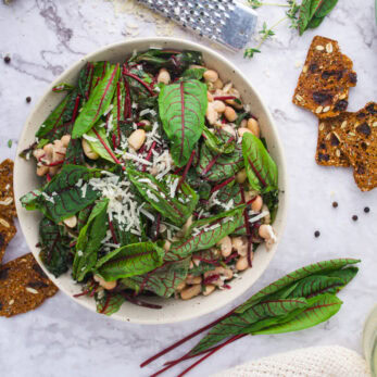 A bowl of Wilted Sorrel and Cannellini Beans sits on a marble surface, surrounded by crispy crackers, fresh greens, black pepper, and a grater.