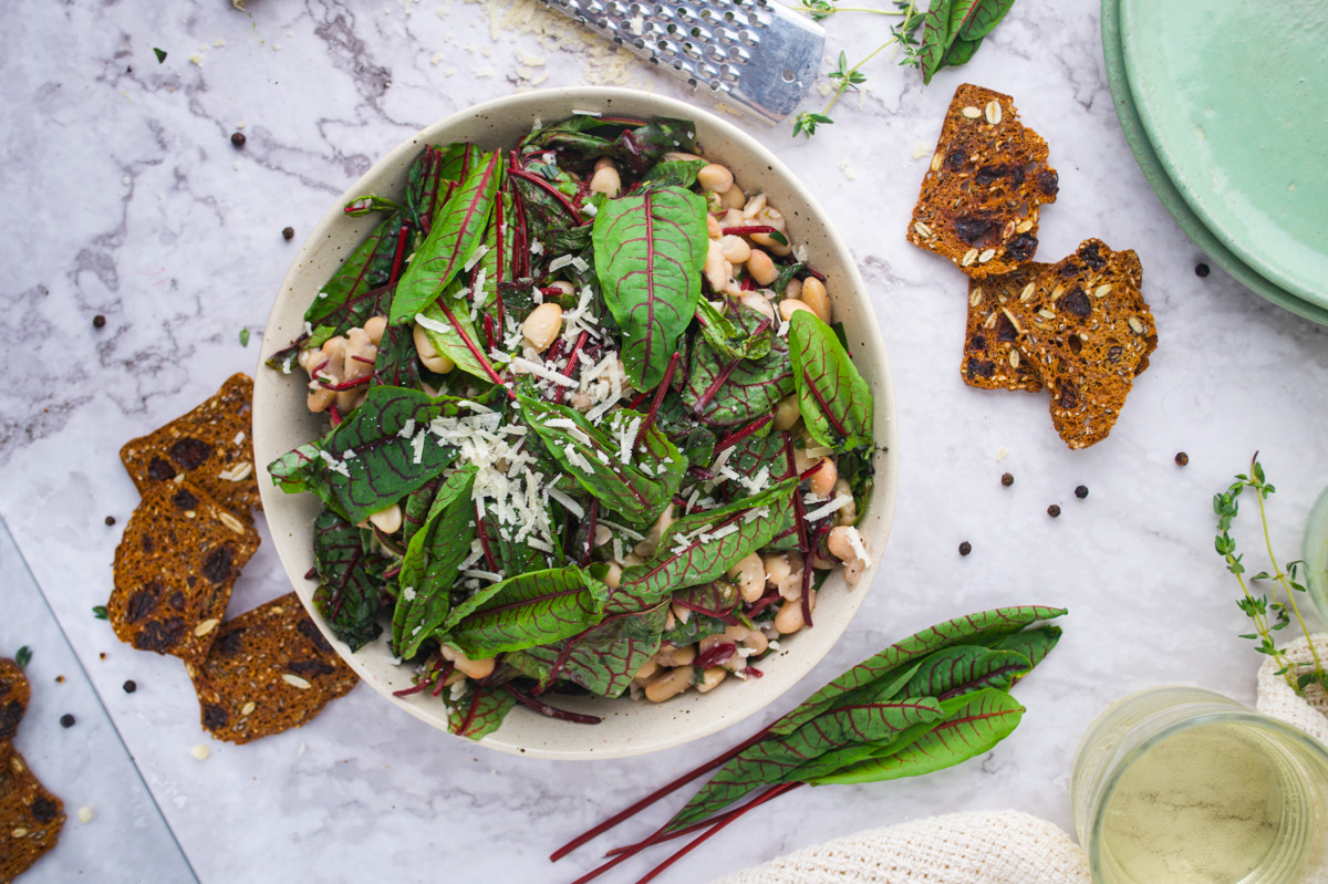 A bowl of Wilted Sorrel and Cannellini Beans sits on a marble surface, surrounded by crispy crackers, fresh greens, black pepper, and a grater.
