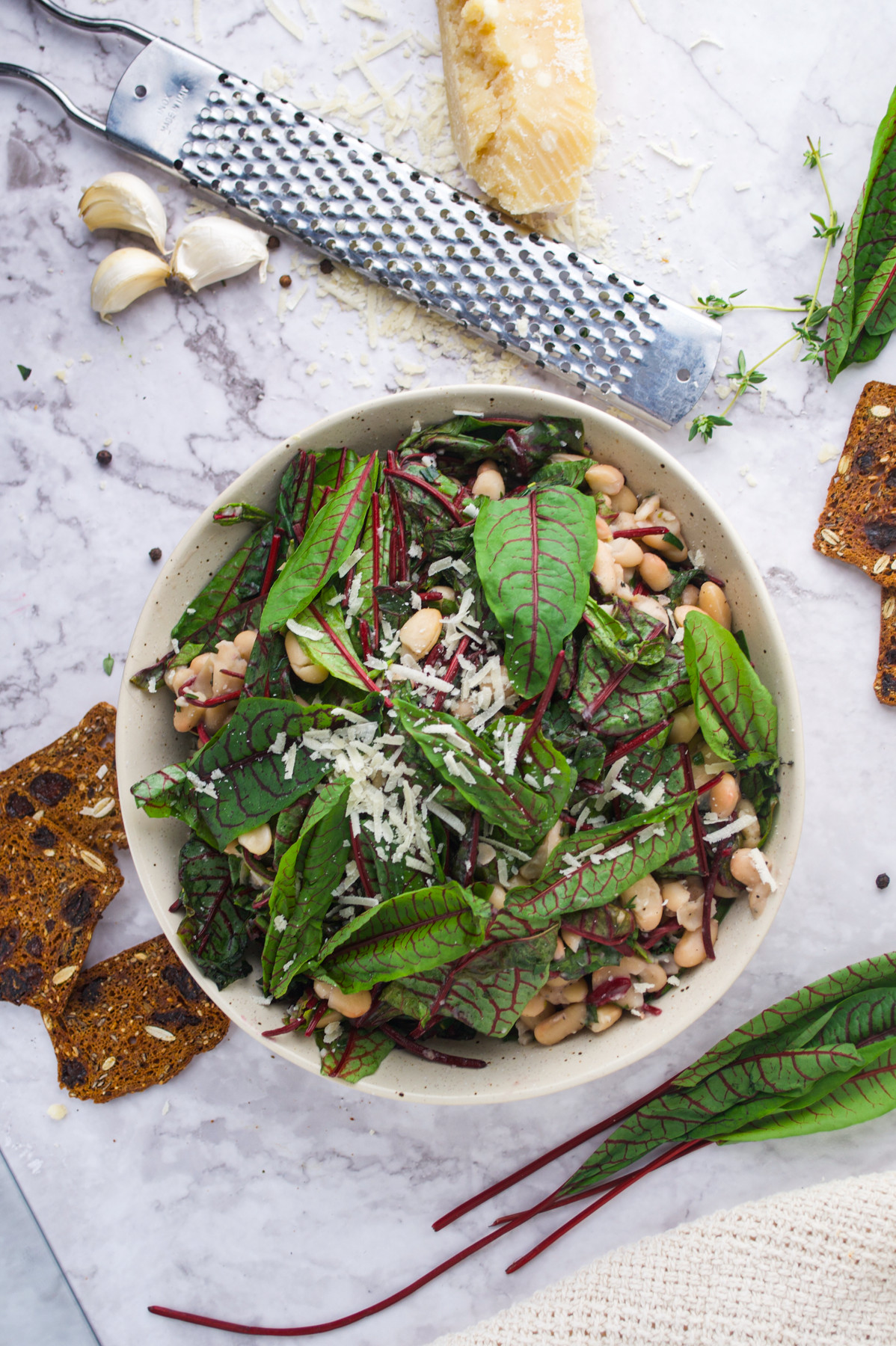 A bowl of Wilted Sorrel and Cannellini Beans surrounded by crackers, a grater, a wedge of cheese, and garlic cloves on a marble surface.