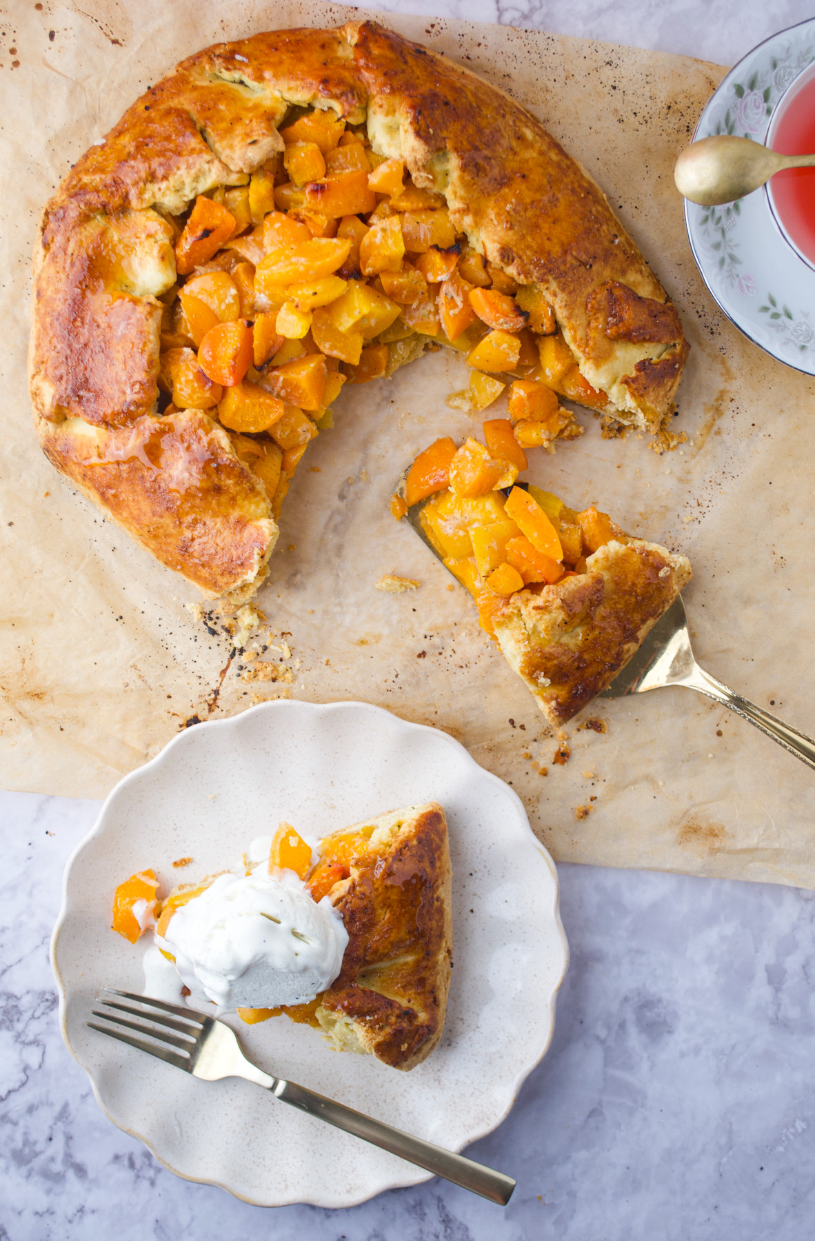 A rustic peach galette with a golden crust sits on parchment paper. A slice is served on a plate, topped with a dollop of whipped cream, next to a fork. A cup of tea is partially visible nearby.