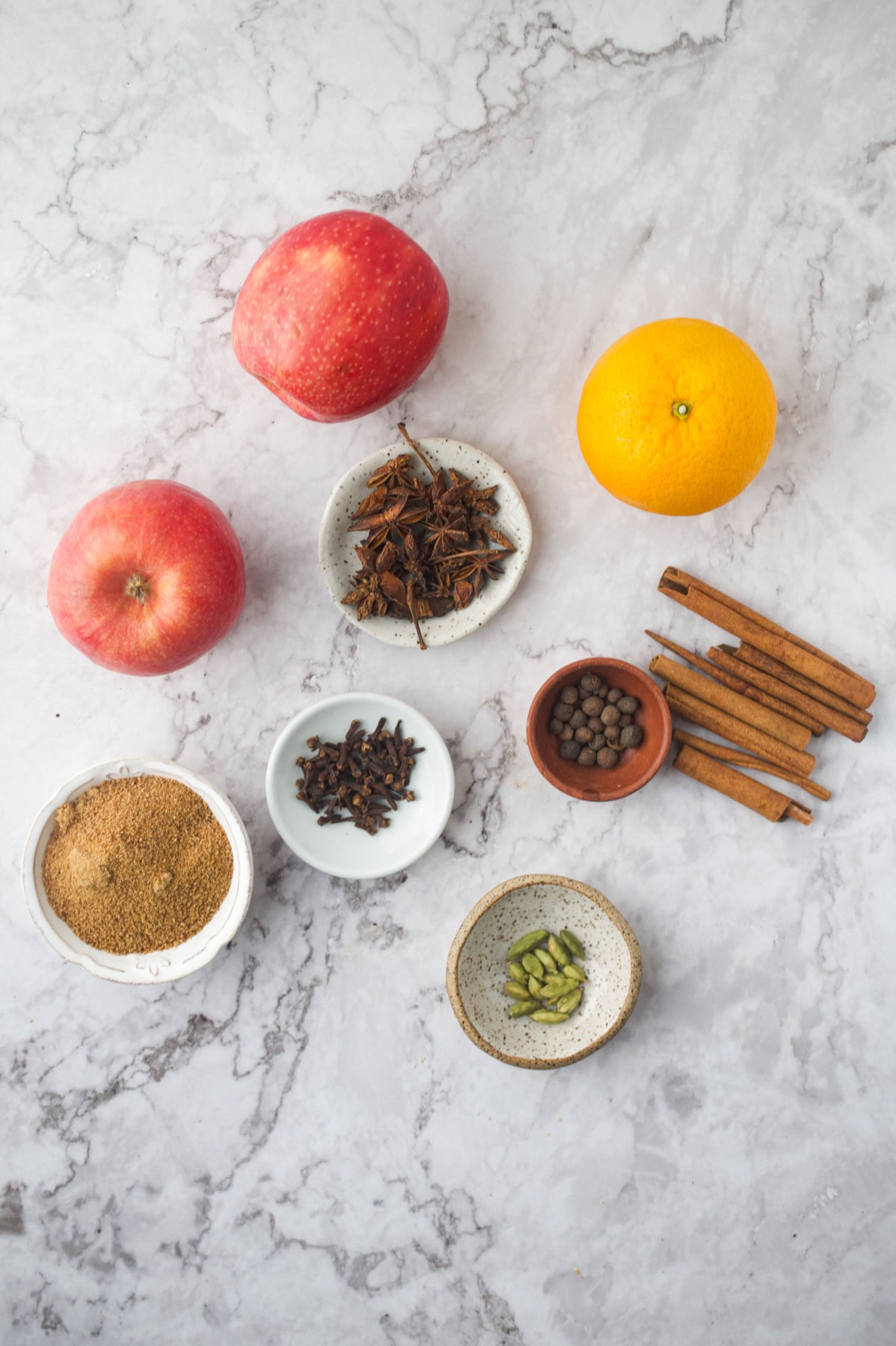 Three apples, one orange, and small bowls containing star anise, whole cloves, allspice, green cardamom pods, cinnamon sticks, and brown sugar arranged on a white marble surface.