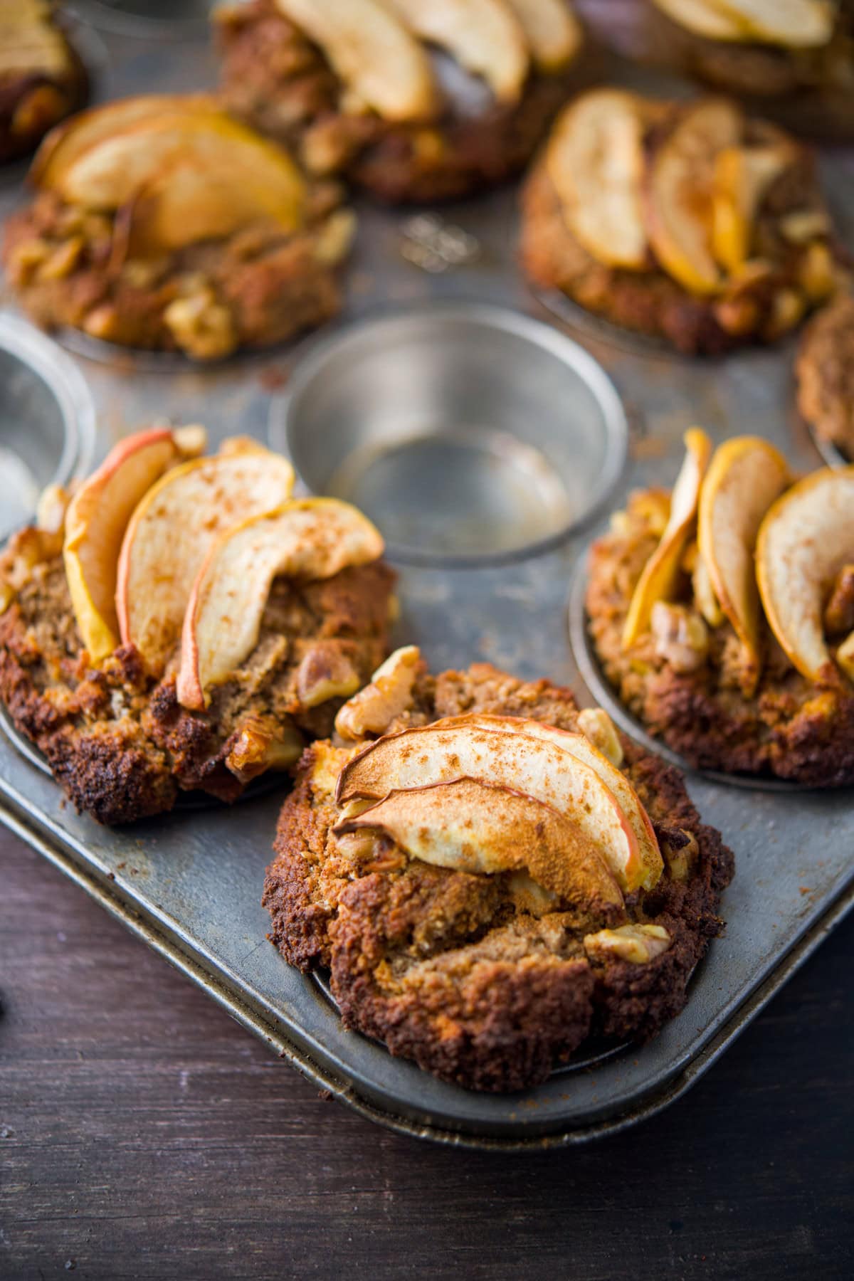 An apple almond muffin tray filled with baked apple muffins, each topped with thin apple slices and a sprinkle of cinnamon, sits on a dark wooden surface. One space in the tray is empty.