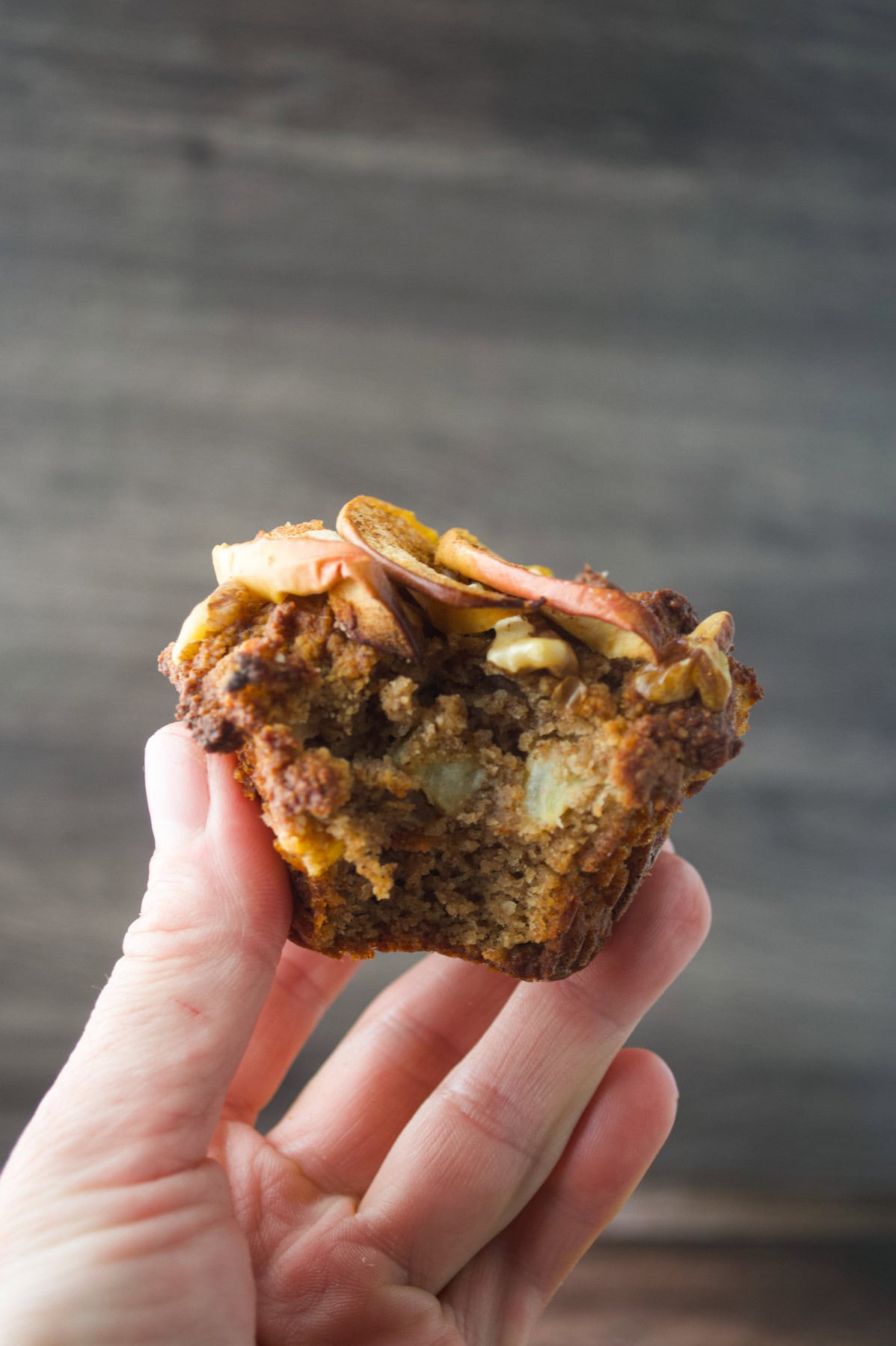 A hand holds up a paleo apple almond muffin with a bite taken out, showing its moist interior. The muffin is topped with thin apple slices and nuts, and is set against a blurred wooden background.