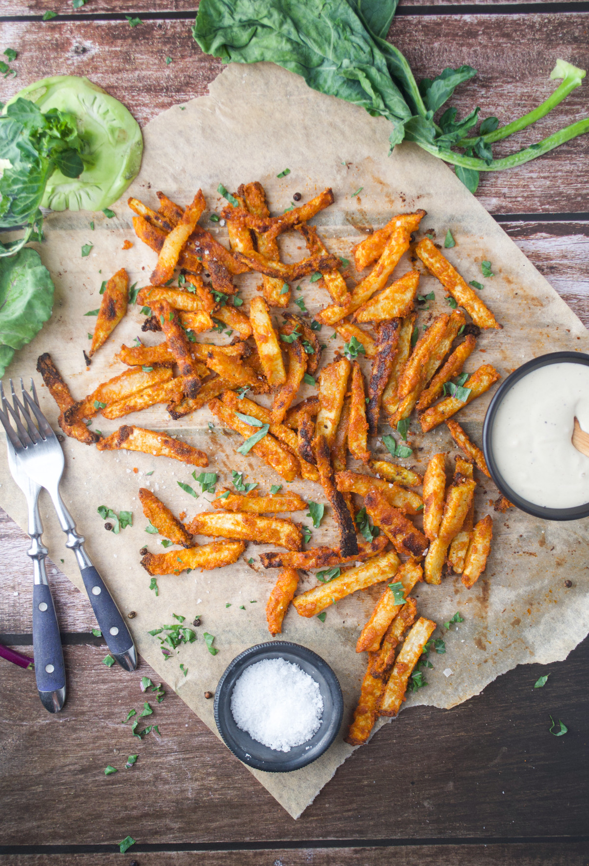 Seasoned French fries on parchment paper, garnished with herbs, next to a bowl of creamy dipping sauce, a small bowl of salt, two forks, and fresh greens on a rustic wooden surface.