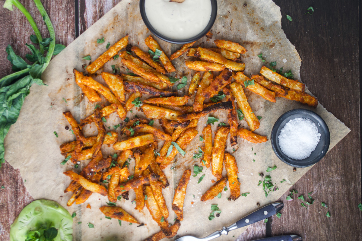Seasoned fries scattered on parchment paper, garnished with chopped herbs. A bowl of white dipping sauce and a small dish of salt are placed nearby, with a fork and knife at the side. Leafy greens are partially visible.