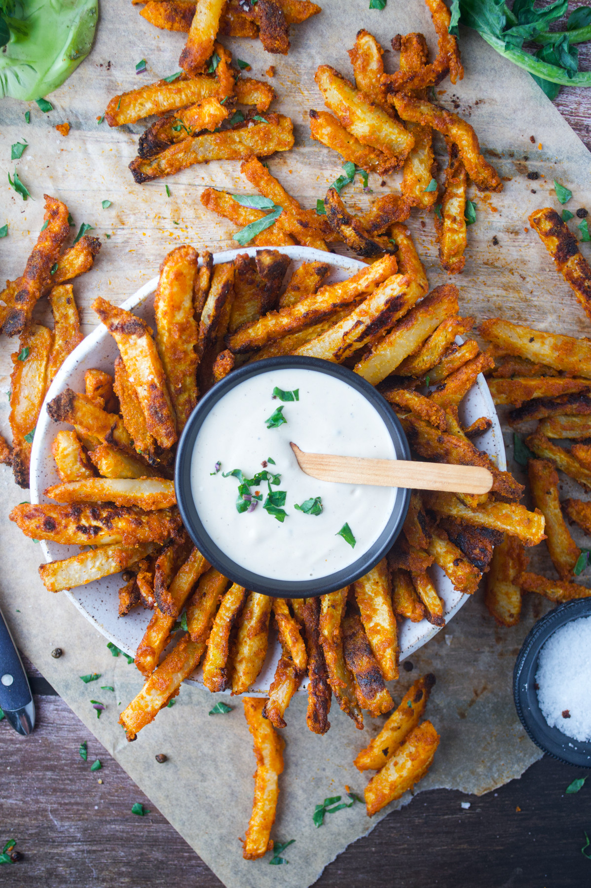 A plate of crispy fries surrounds a bowl of white dipping sauce garnished with herbs, with fries scattered on parchment paper and a salt dish nearby.