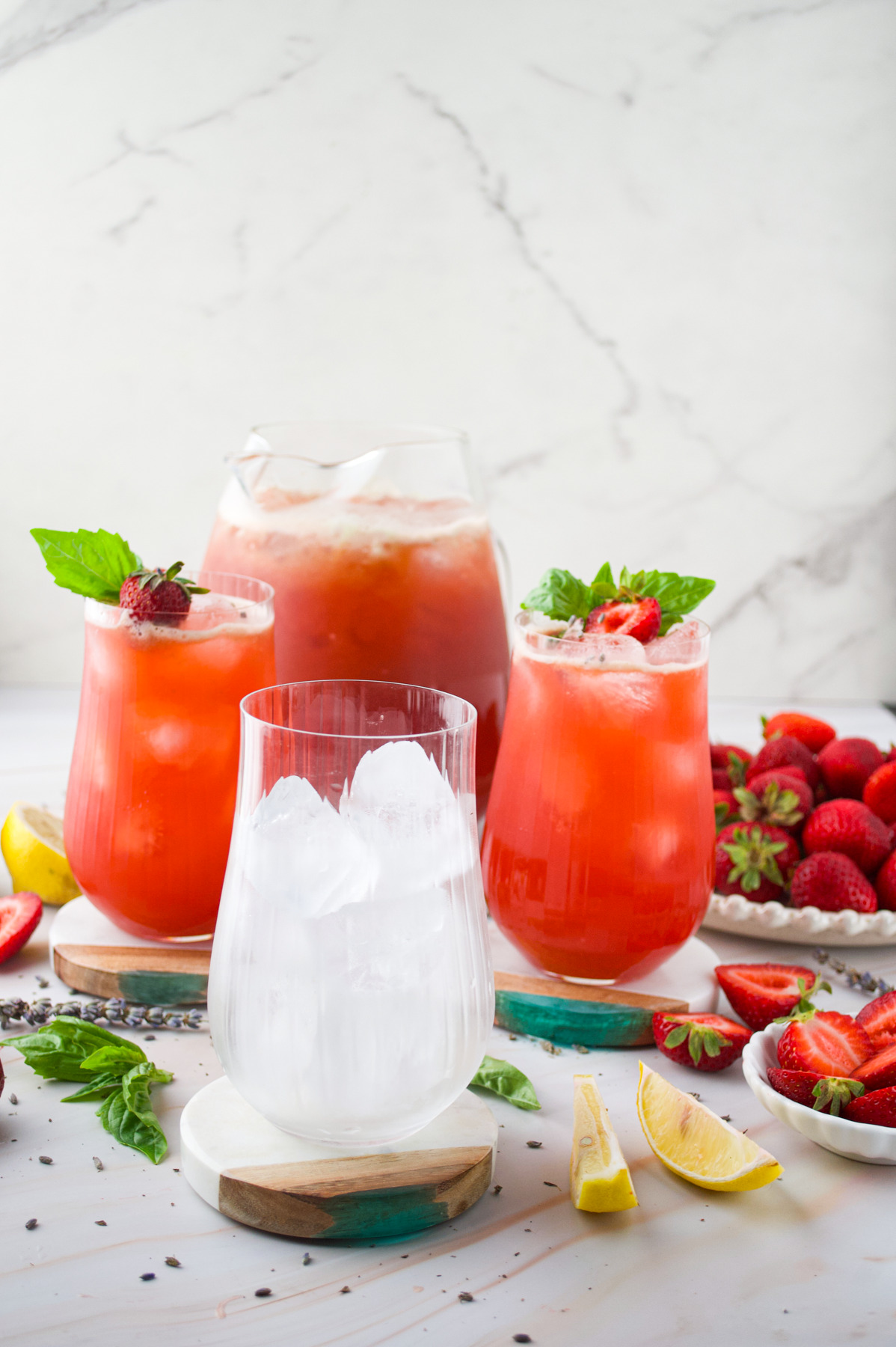 Three glasses of vibrant Strawberry Lavender Lemonade, garnished with basil and strawberries, sit on a white surface with an empty glass of ice in front. Fresh strawberries, lemon wedges, and basil leaves are scattered around.
