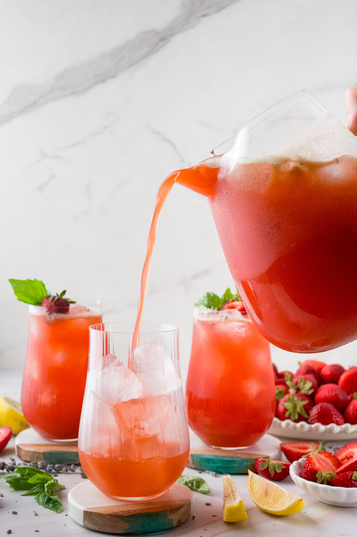 A hand pours a bright red, Strawberry Lavender Lemonade from a pitcher into a glass. Two more filled glasses, strawberries, lemon wedges, and mint leaves sit on a light surface in the background.