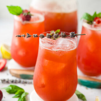 Four glasses of Strawberry Lavender Lemonade garnished with herbs and berries, surrounded by fresh ingredients like basil leaves, lemon wedges, and spices on a white surface. The drinks look refreshing and summery.