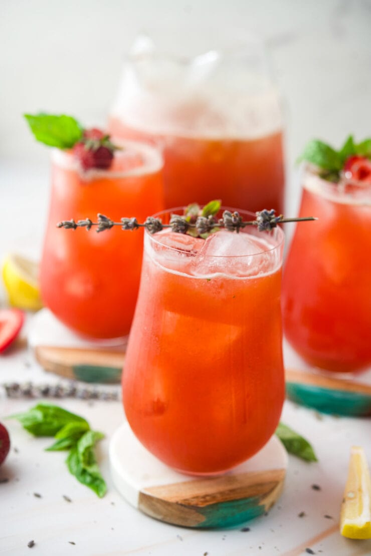 Four glasses of Strawberry Lavender Lemonade garnished with herbs and berries, surrounded by fresh ingredients like basil leaves, lemon wedges, and spices on a white surface. The drinks look refreshing and summery.