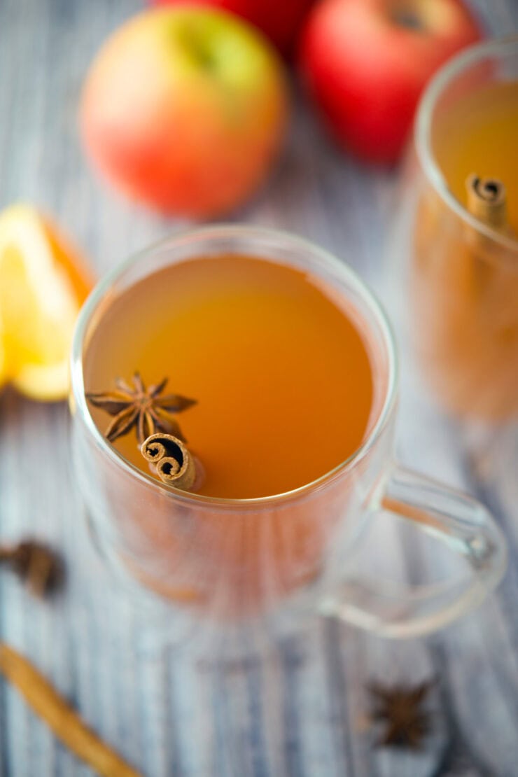 A clear glass mug filled with spiced apple cider, garnished with a cinnamon stick and star anise. Out of focus apples and an orange slice are in the background on a striped surface.