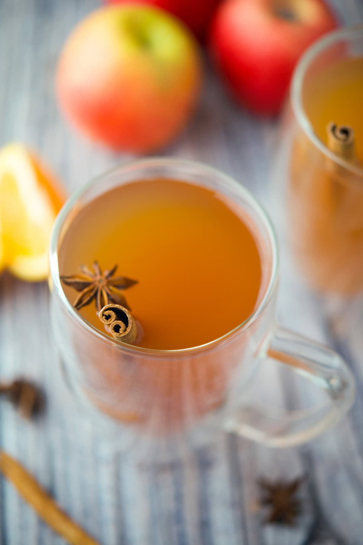 A clear glass mug filled with spiced apple cider, garnished with a cinnamon stick and star anise. Out of focus apples and an orange slice are in the background on a striped surface.