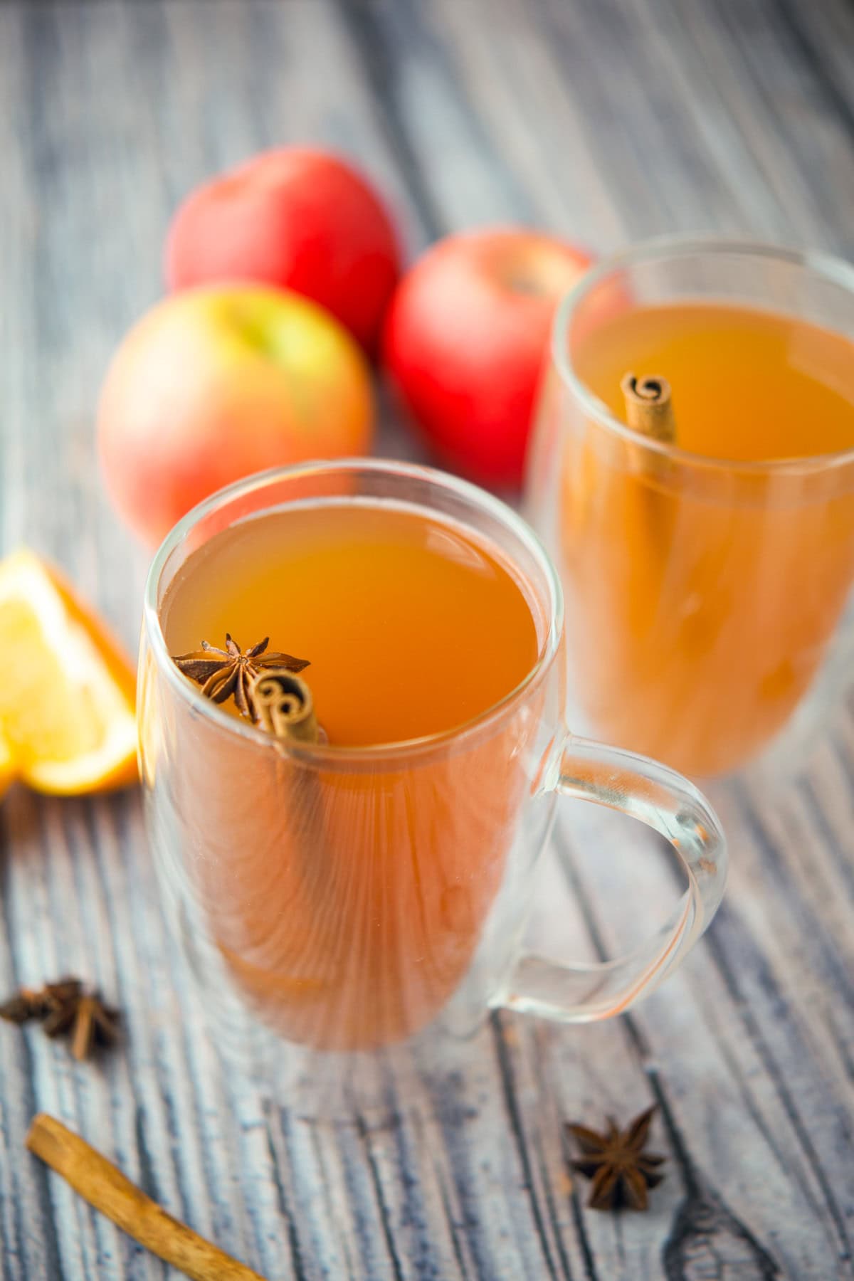 Two clear glass mugs filled with spiced apple cider, each garnished with a cinnamon stick and star anise. Three apples and an orange wedge are in the background on a wooden surface.