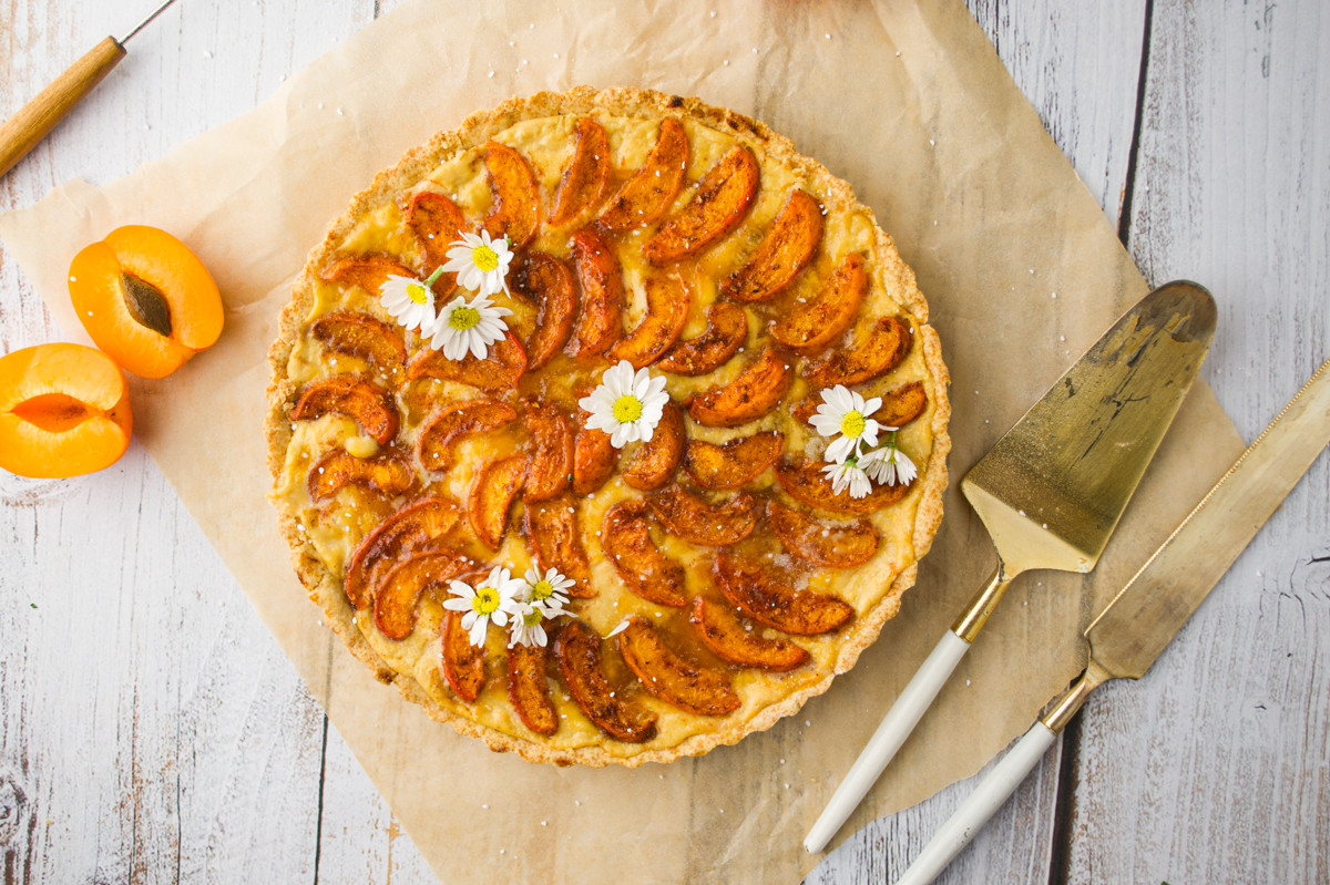Apricot tart topped with fresh apricot slices and small white flowers, placed on parchment paper on a rustic white wooden surface. Two apricot halves and gold serving utensils are beside the tart.