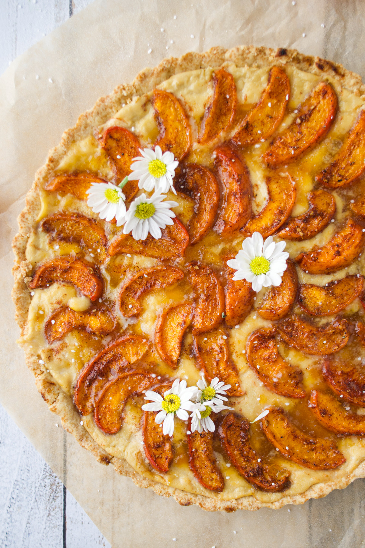 An Apricot Tart with Custard (Gluten-Free), garnished with three white daisy flowers, sits on parchment paper on a wooden surface.