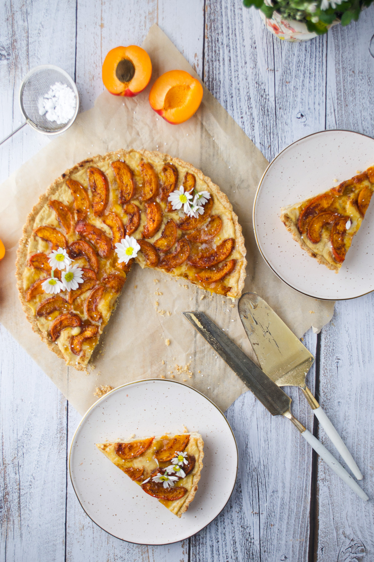 A rustic apricot tart garnished with white flowers is on parchment paper, with two slices served on plates nearby. Fresh apricots, a cake server, and a knife are arranged on a white wooden surface.