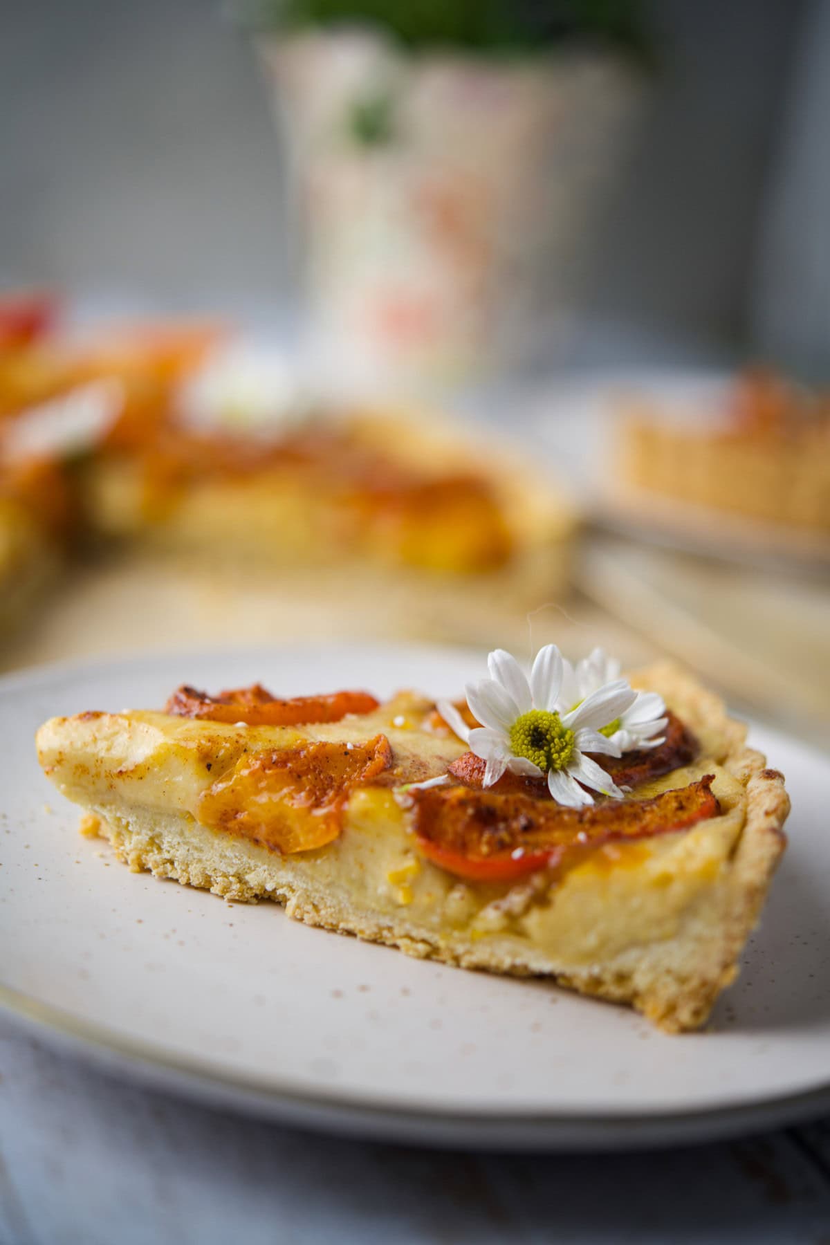 A slice of tart with golden crust and orange toppings is served on a plate, garnished with a small white daisy. Blurred background shows more tart slices and a floral-patterned object.