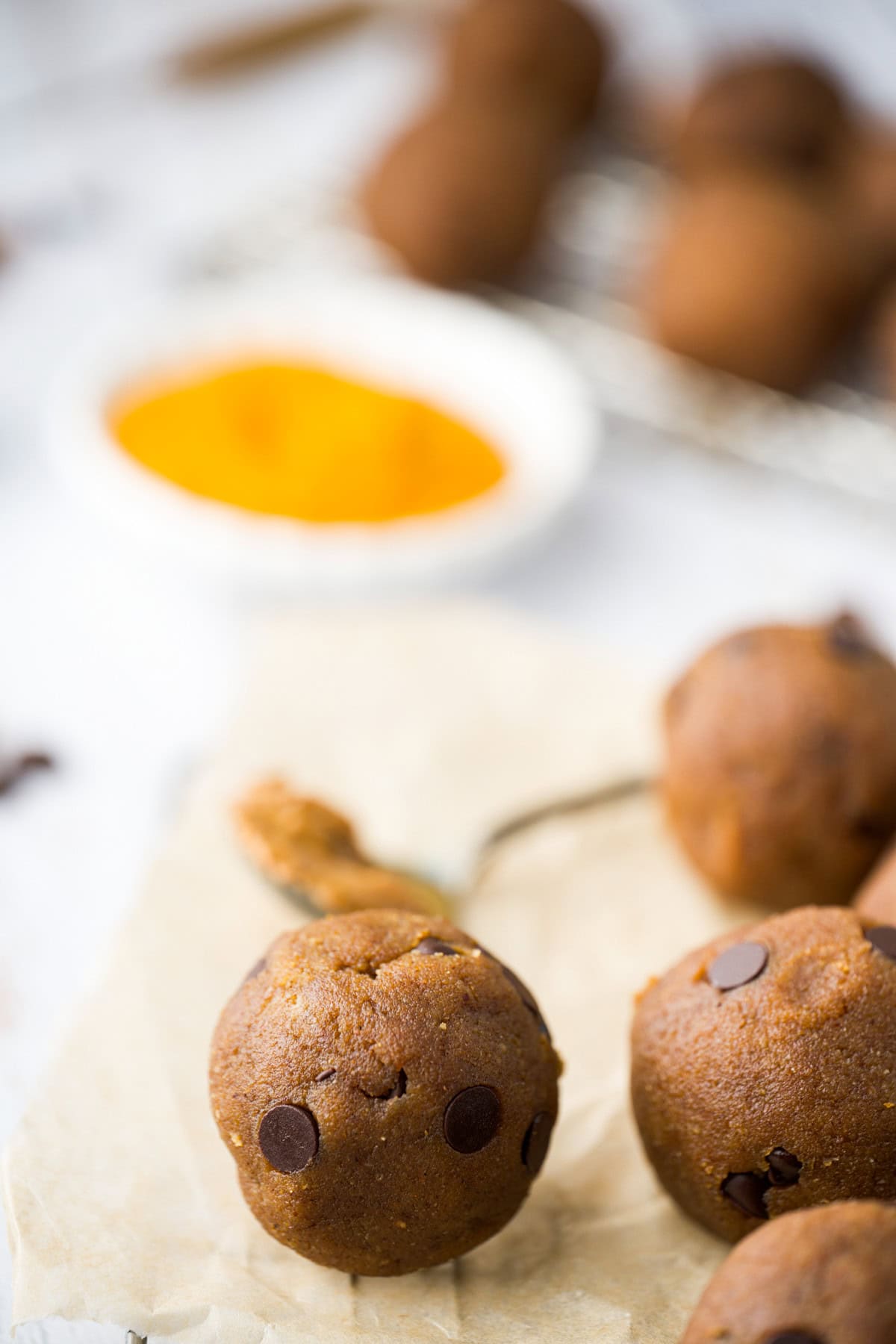 A close-up of round, brown cookie dough balls with chocolate chips on parchment paper. In the blurred background, there is a small bowl of yellow powder and more dough balls.
