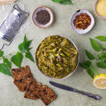 A bowl of nettle pistachio pesto with pistachios, surrounded by crackers, nettle leaves, half a lemon, spices, a cheese grater, and a knife on a light green surface.