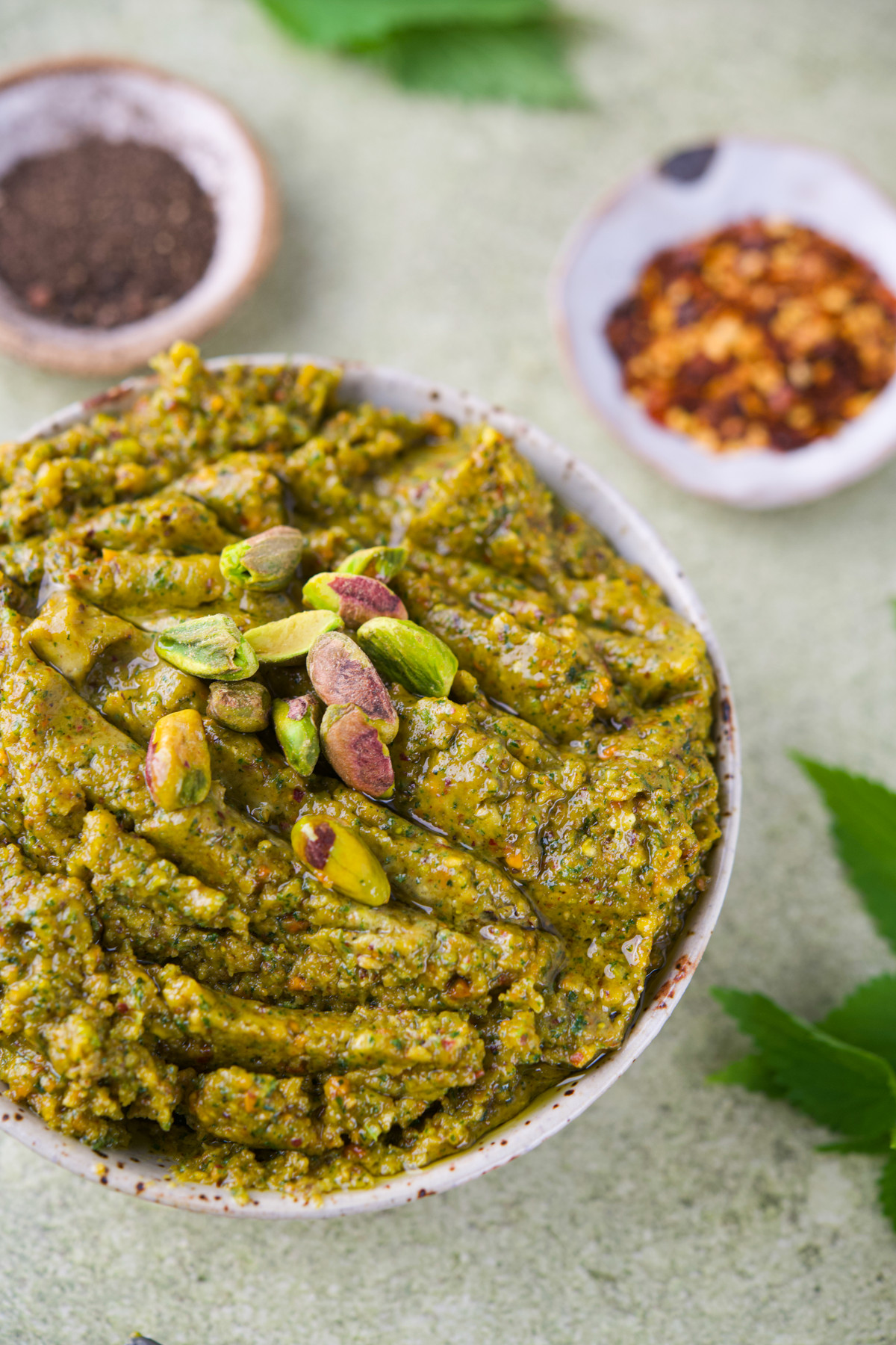 A bowl of nettle pistachio pesto garnished with whole pistachios, with dishes of black pepper and red pepper flakes and green leaves in the background.