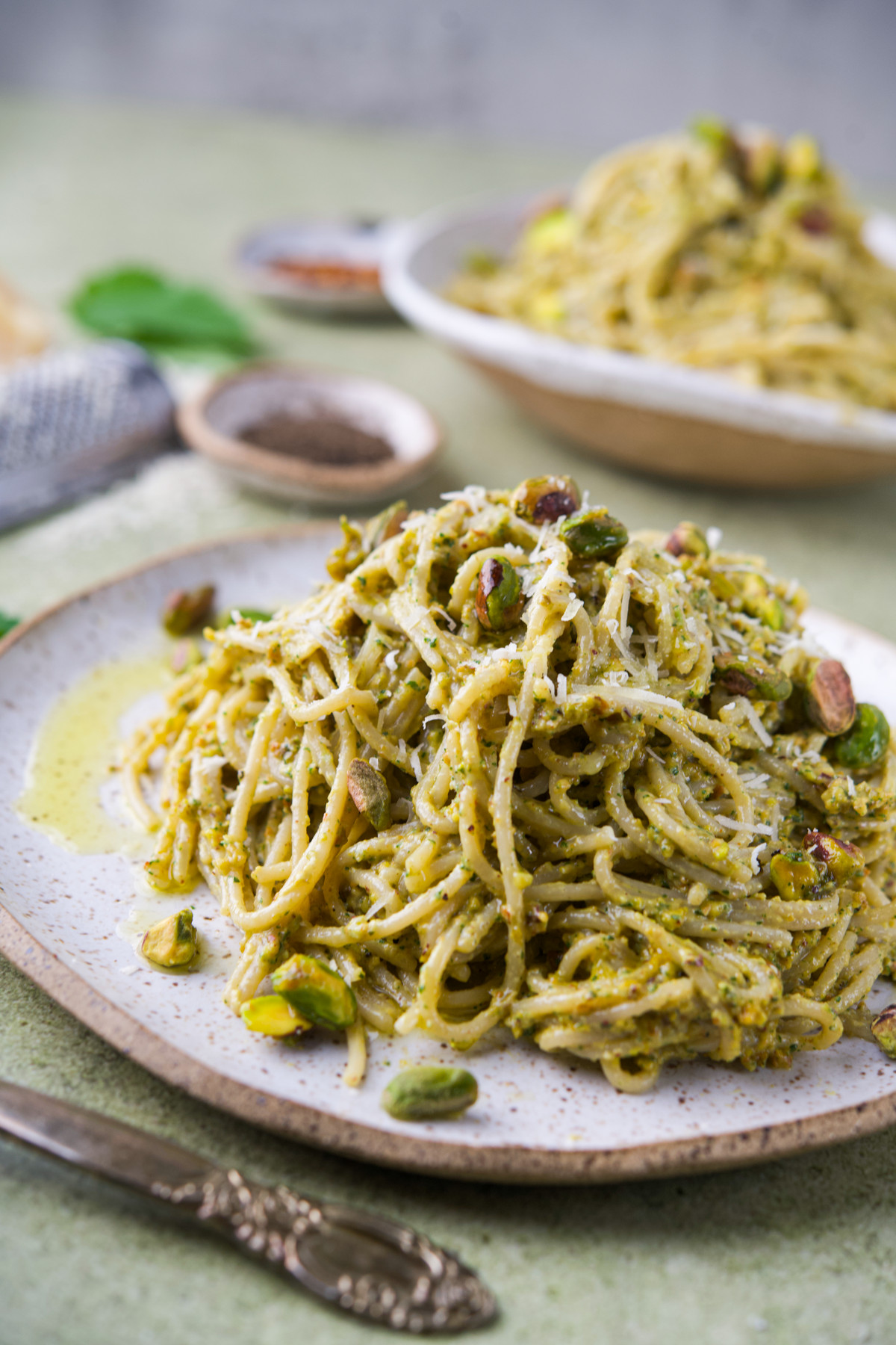 A plate of spaghetti coated in nettle pistachio pesto garnished with grated cheese and whole pistachios, sits on a ceramic plate with a fork beside it. Another plate of spaghetti is blurred in the background.