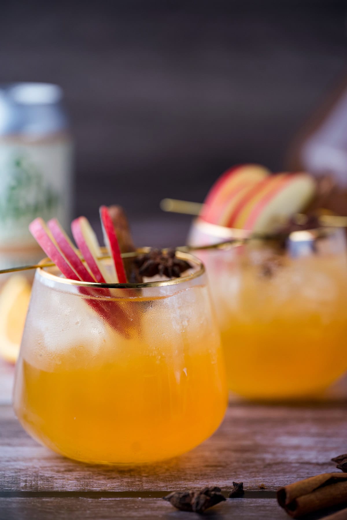 Two glasses of Hard Apple Cider Ginger Spritzer with ice, topped with sliced red apples and a star anise, sit on a wooden surface. A blurred drink can and bottle are visible in the background.