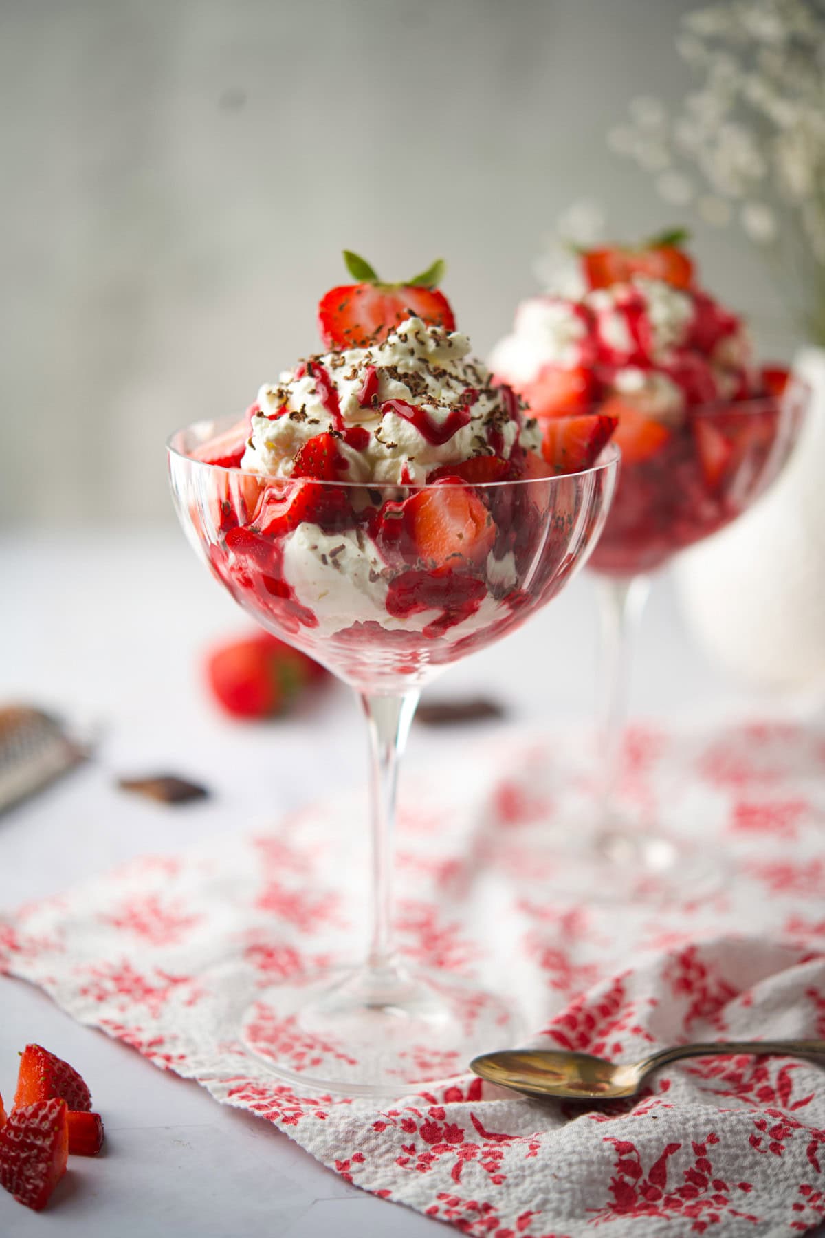 Two elegant glass dishes filled with layers of whipped cream, fresh strawberries, and chocolate shavings, set on a red-patterned napkin with a spoon and strawberries in the background.