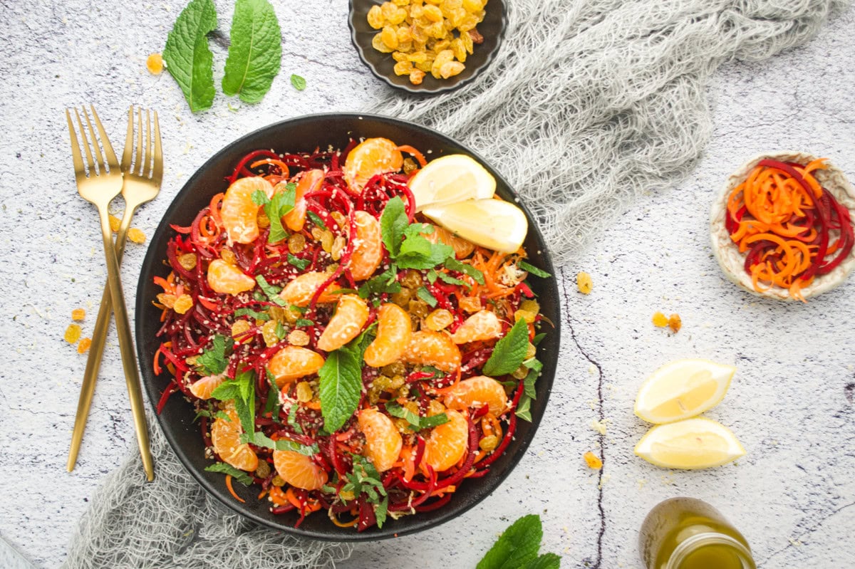 A colorful salad with mandarin orange segments, spiralized beets and carrots, golden raisins, mint leaves, and seeds in a black bowl, garnished with lemon wedges, surrounded by forks, extra fruit, and herbs on a light textured surface.