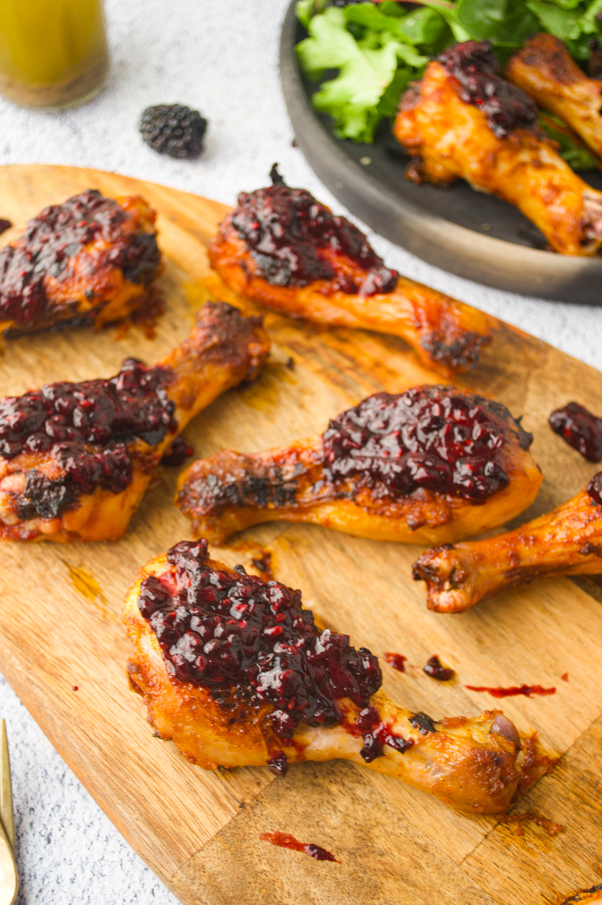 Grilled chicken drumsticks topped with a glossy dark berry sauce are arranged on a wooden cutting board, with a plate of greens and more drumsticks visible in the background.