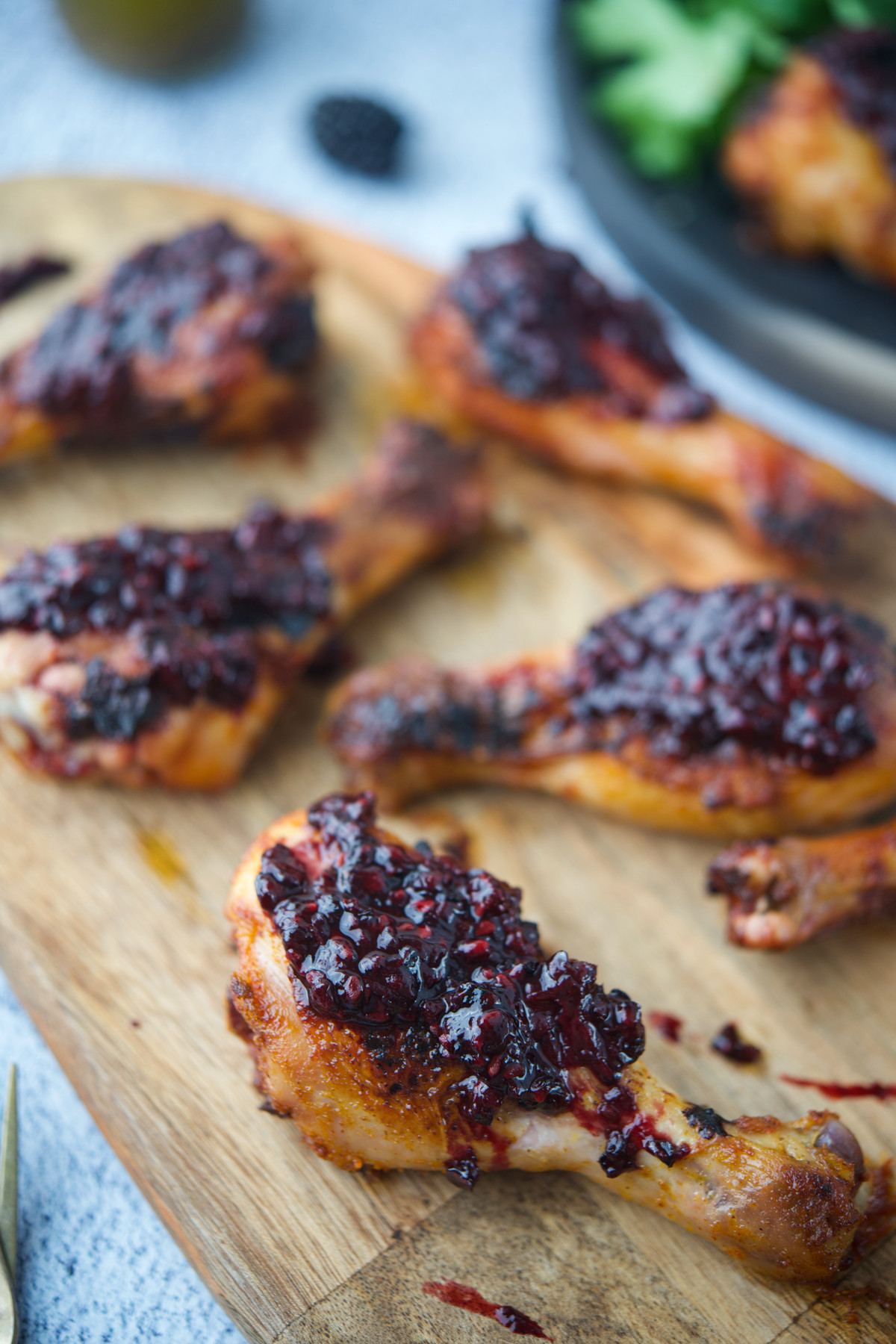 Roasted chicken drumsticks on a wooden board, topped with a chunky, dark berry sauce. More chicken and leafy greens are visible blurred in the background.