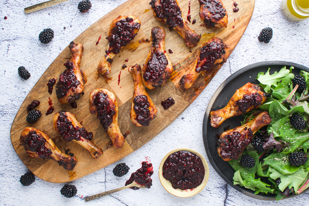 Grilled chicken drumsticks topped with blackberry sauce on a wooden board, with fresh blackberries scattered around. A black plate with salad greens and drumsticks, and a small bowl of blackberry sauce are nearby.
