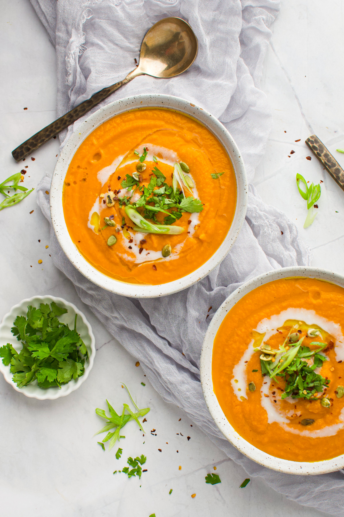 Two bowls of creamy carrot ginger soup garnished with cream, sliced green onions, seeds, and herbs sit on a light cloth with spoons and a small dish of fresh cilantro beside them.