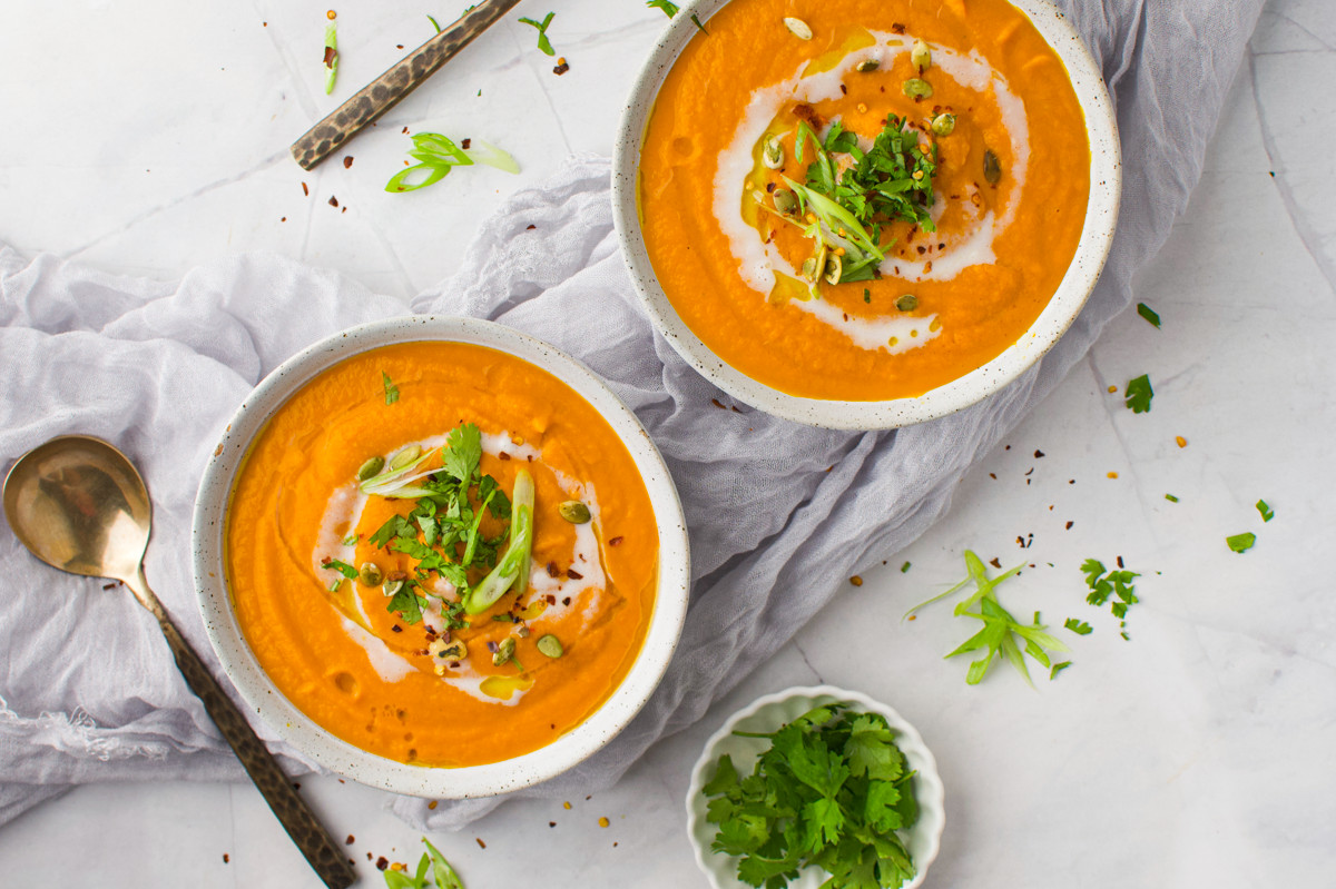 Two bowls of creamy orange soup topped with sliced green onions, herbs, and seeds on a light cloth, with a spoon and chopped parsley on the side.