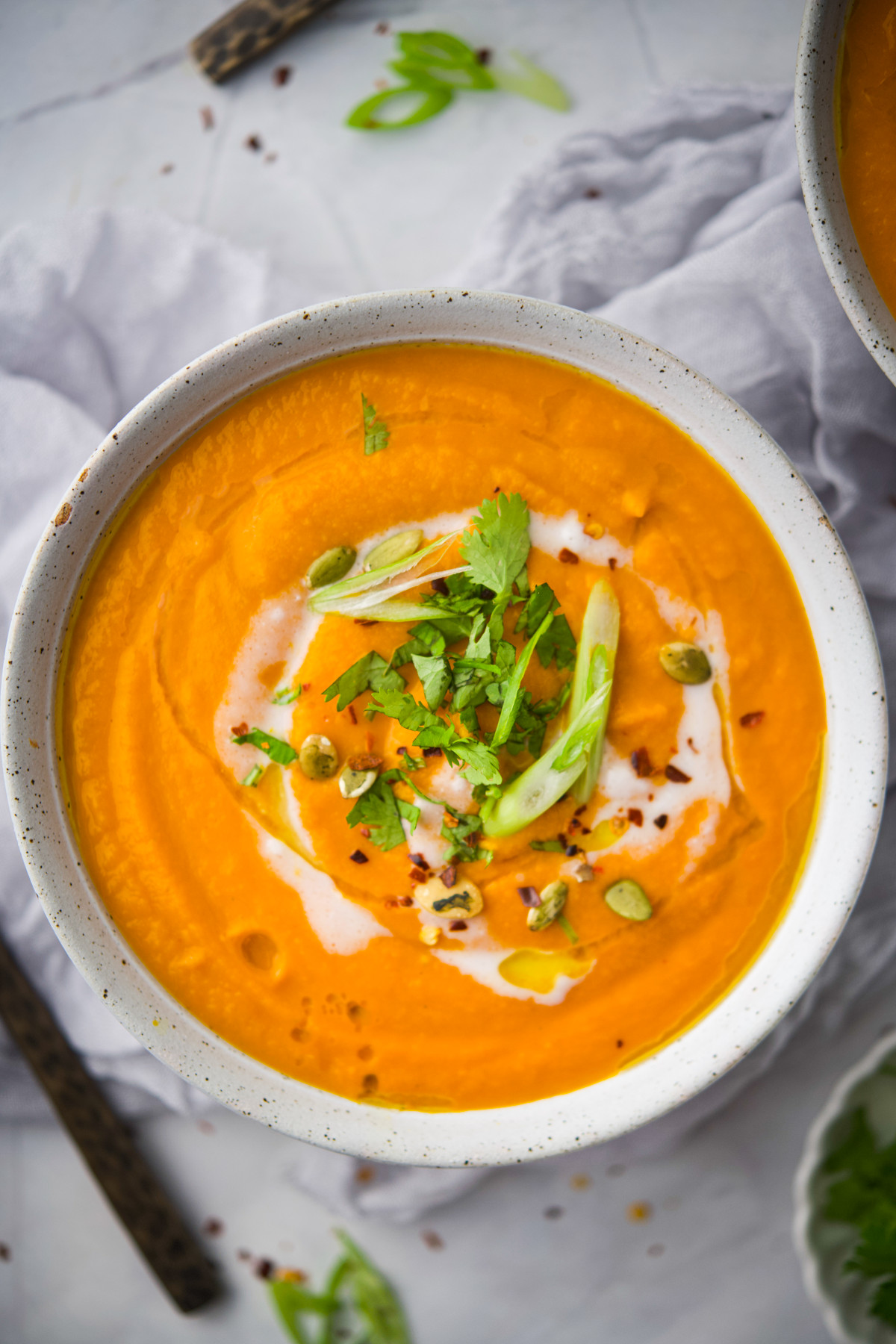 A bowl of creamy orange soup garnished with green herbs, sliced scallion, seeds, red pepper flakes, and a swirl of cream, on a light cloth background.
