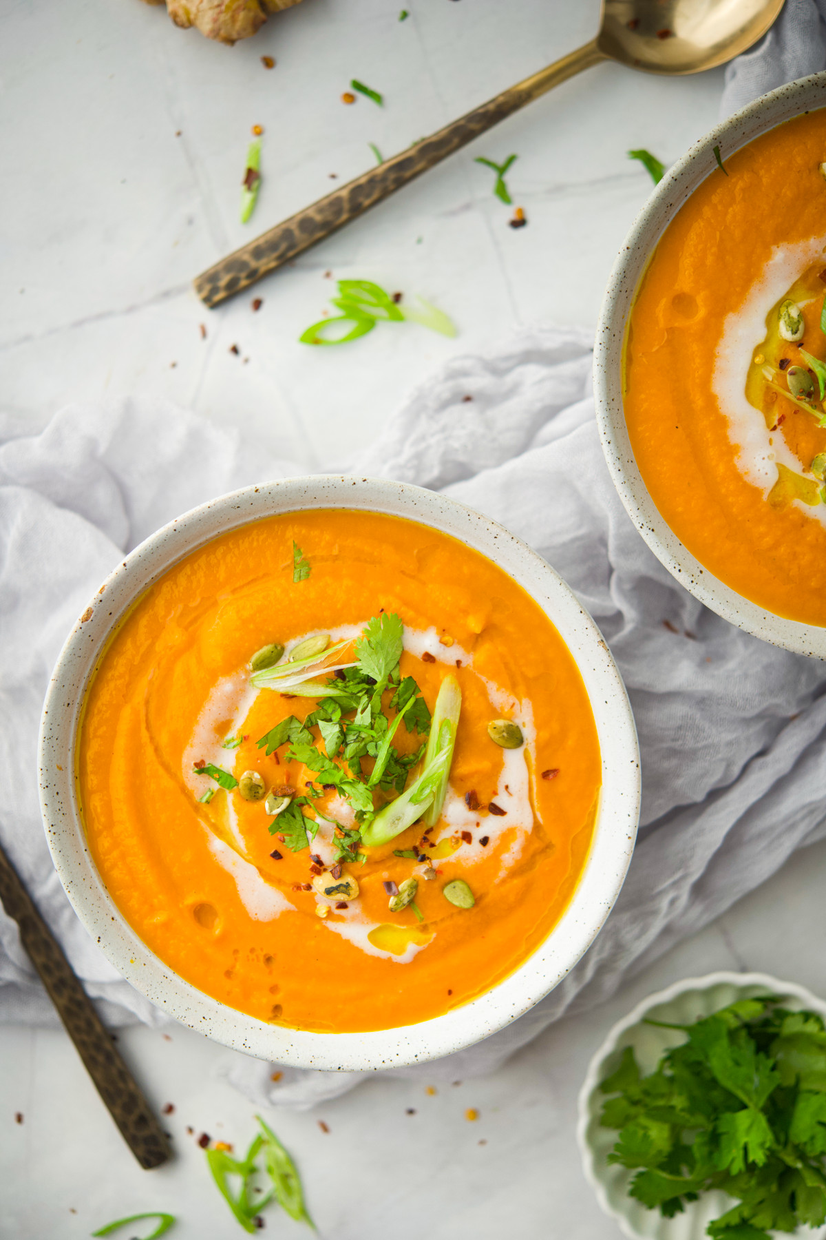 Two bowls of Creamy Carrot Ginger Soup garnished with sliced green onions, cilantro, pumpkin seeds, and a swirl of cream on a white surface with a gray cloth, spoon, and herbs nearby.