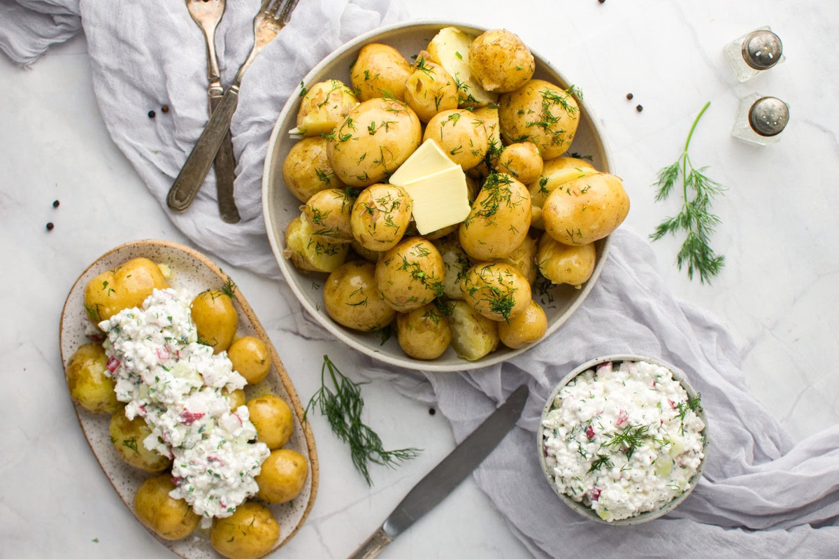 A bowl of boiled new potatoes garnished with dill and a pat of butter, a plate of potatoes topped with a creamy salad, and a bowl of the same salad, all on a white surface with herbs and utensils.