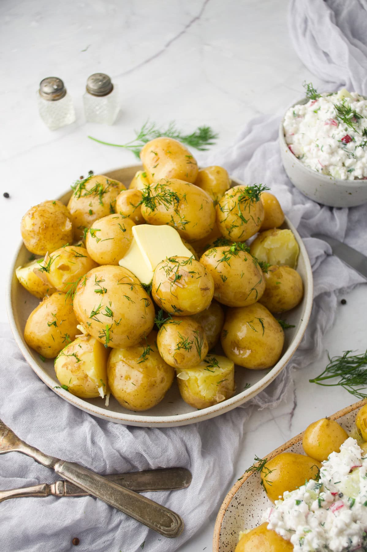 A bowl of boiled baby potatoes topped with fresh dill and pats of butter, surrounded by cutlery, a bowl of creamy salad with herbs, and salt and pepper shakers on a light linen cloth.