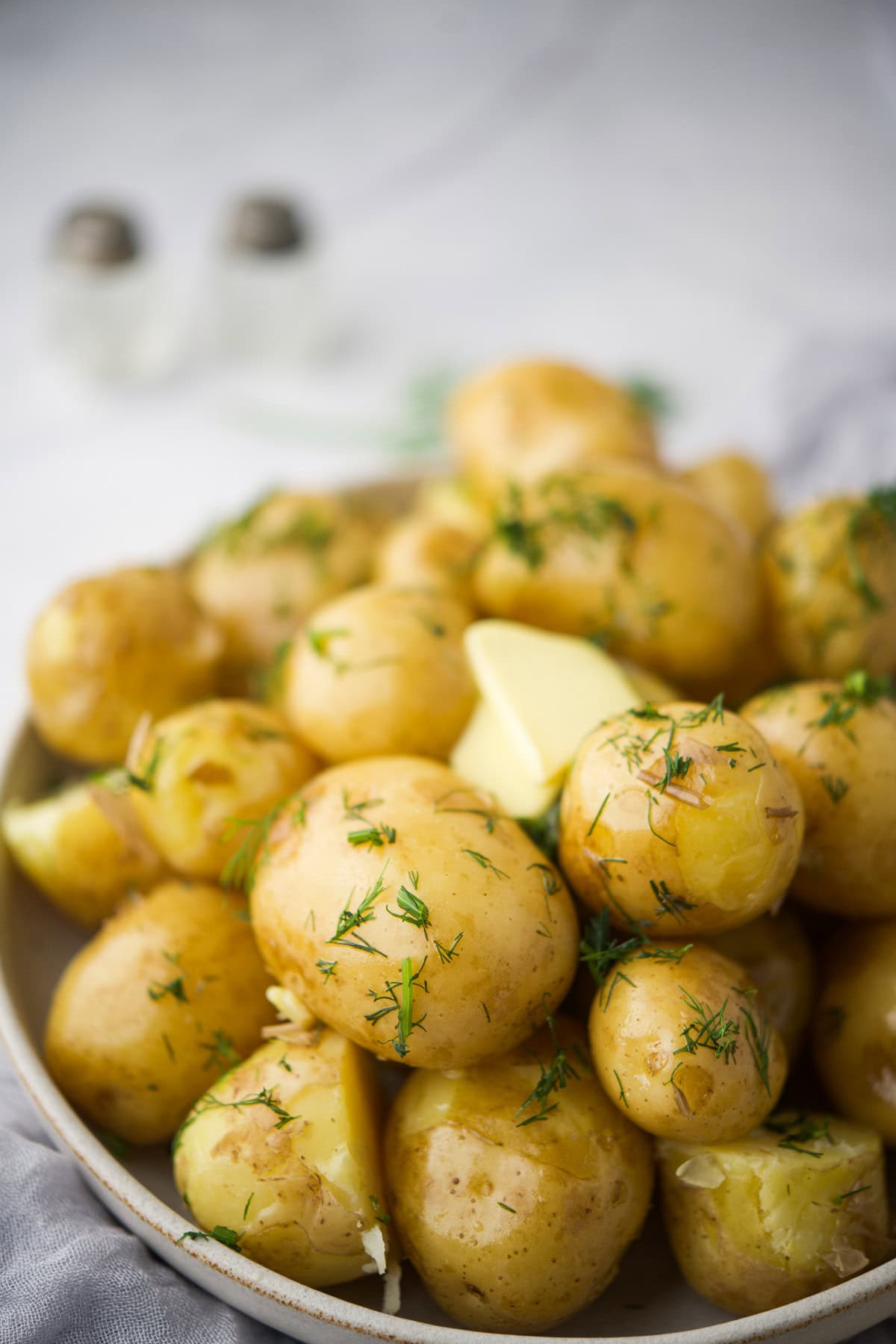 A close-up of a plate filled with boiled baby potatoes, garnished with fresh chopped dill and a pat of butter. Salt and pepper shakers are blurred in the background.