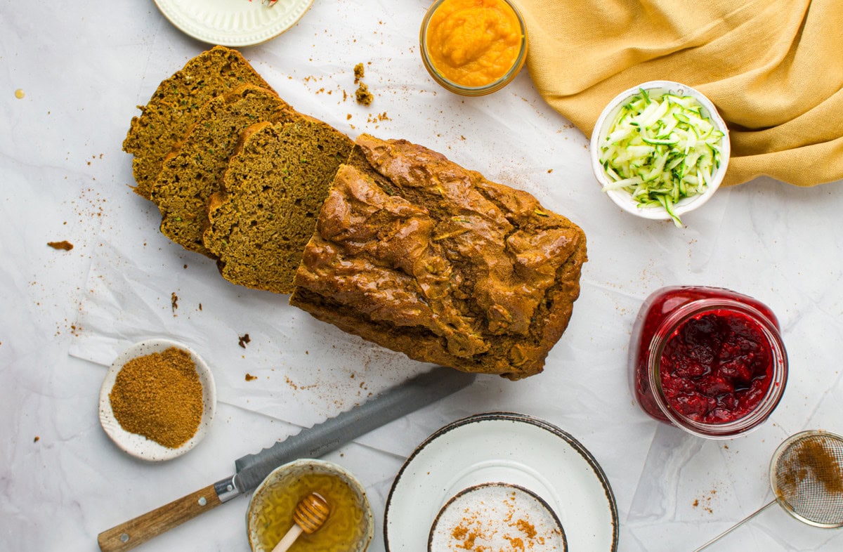 A loaf of bread is sliced on a white surface, surrounded by bowls of shredded zucchini, fruit preserves, brown sugar, honey, and a jar of orange puree, with a knife and plates nearby.