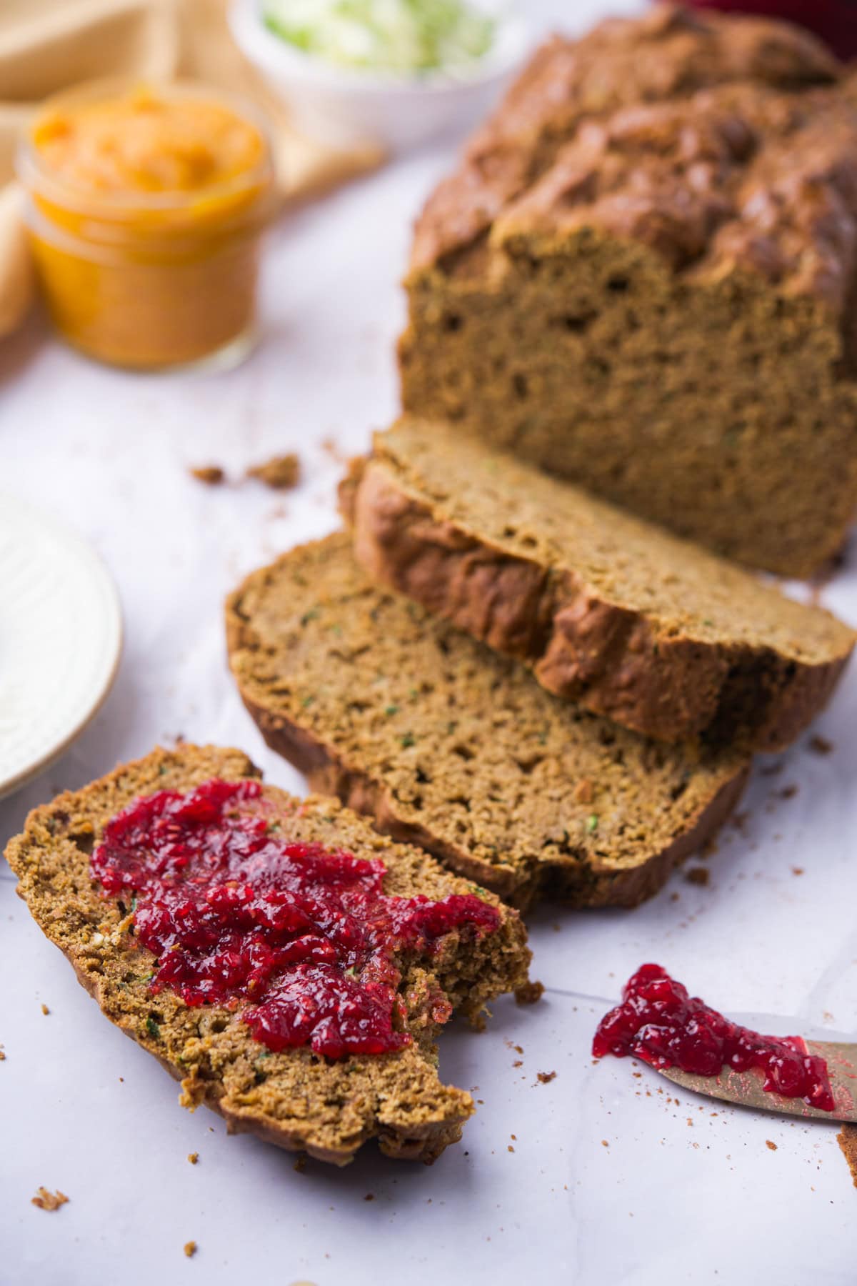 Sliced brown bread on a white surface with one slice topped with red berry jam. A jar with orange spread and a bowl with green food are blurred in the background.