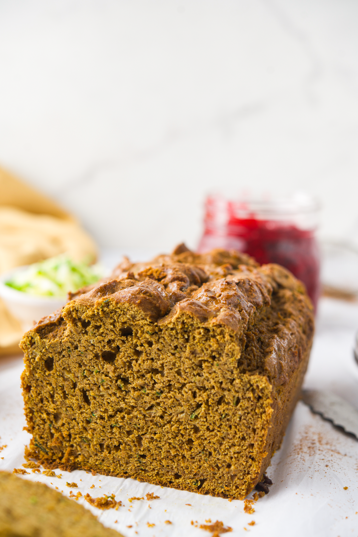 A close-up of a sliced loaf of zucchini bread on parchment paper, with a jar of red jam and a small bowl of shredded zucchini in the blurred background.