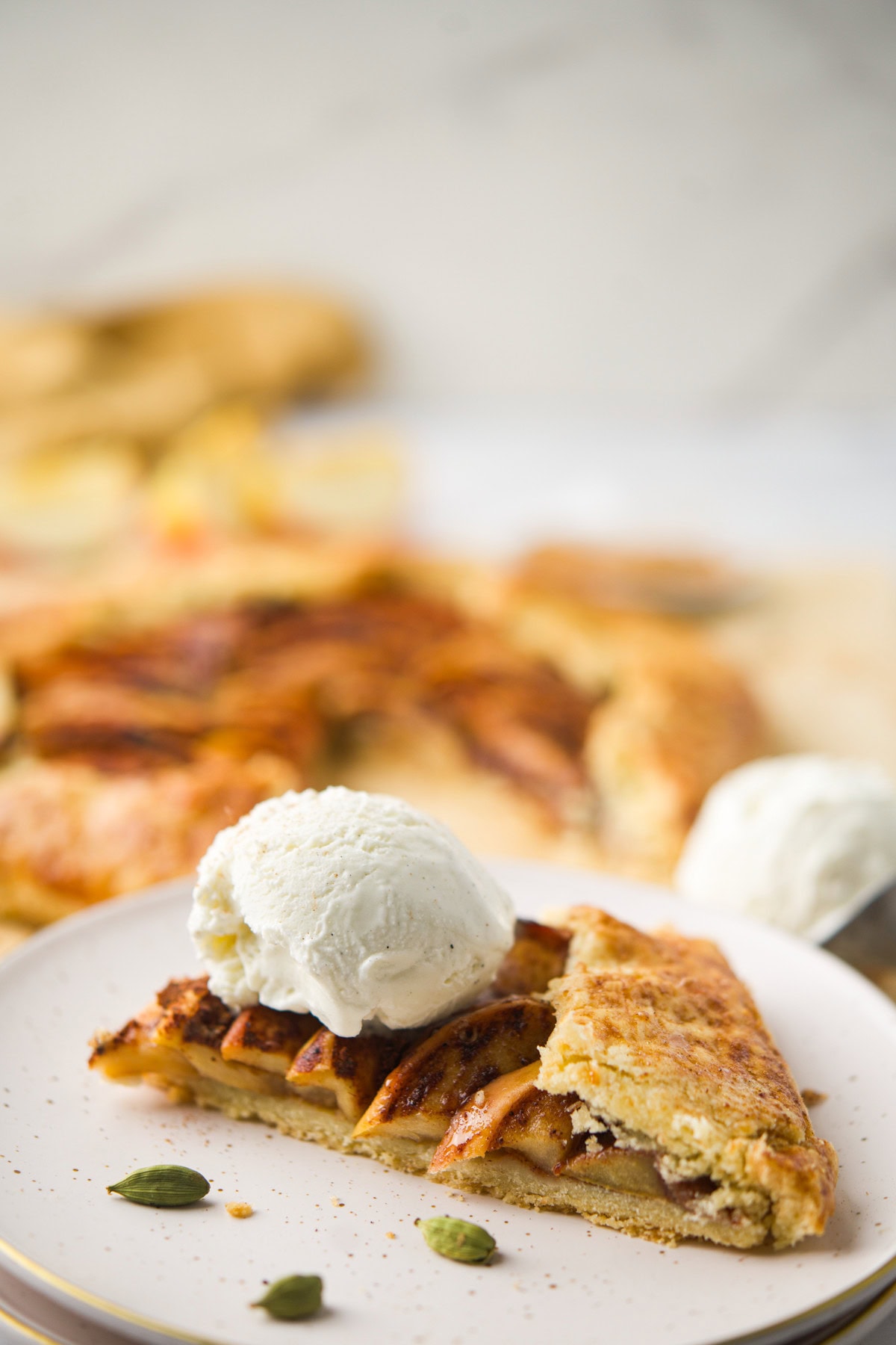 A slice of apple pie topped with a scoop of vanilla ice cream sits on a plate, with whole cardamom pods nearby and more pie visible in the blurred background.