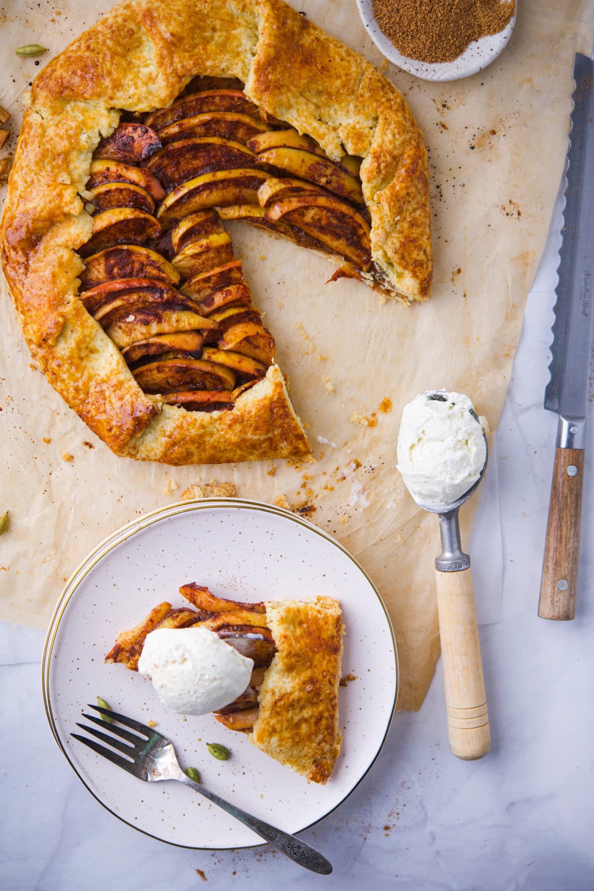 A rustic apple galette with a slice cut out sits on parchment paper. The slice is plated on a dish with a scoop of vanilla ice cream, next to a fork and a knife. A bowl of brown sugar is visible in the corner.